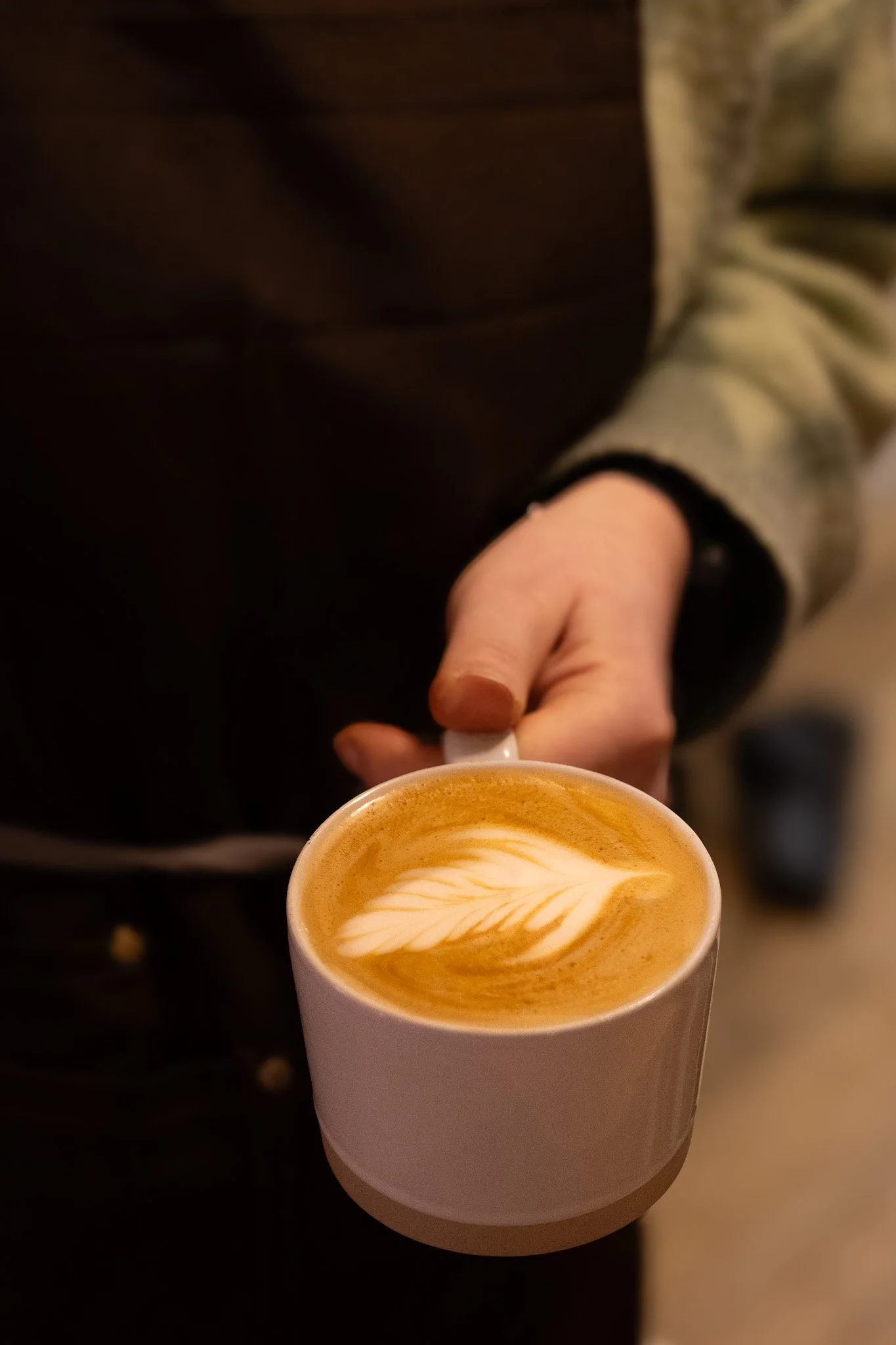 A person holding a white mug of coffee with latte art, featuring a leaf pattern, in a cozy indoor setting. The New England Coffee House.