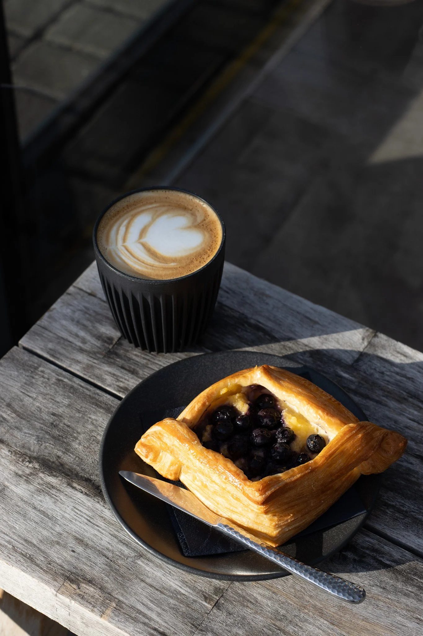 A cup of coffee with latte art and a blueberry Danish pastry on a black plate, placed on a rustic wooden table outdoors. The Scandinavian Coffee Pod.