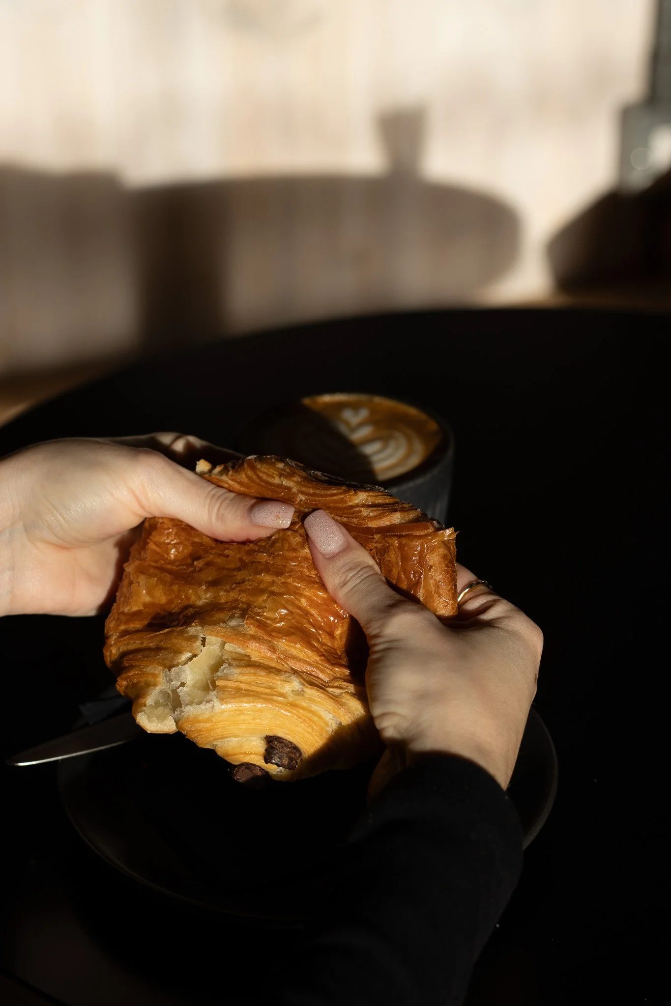 Person holding a piece of a chocolate croissant in front of a coffee cup on a black plate, with a blurred background. The Scandinavian Coffee Pod.