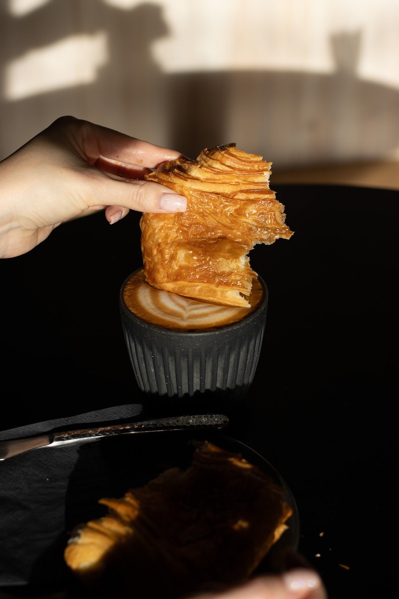 A hand holding a piece of flaky, crispy pastry over a cup of coffee with latte art. The background is blurred. The Scandinavian Coffee Pod.