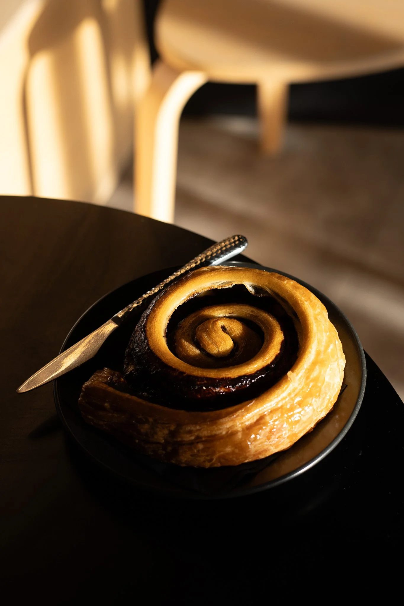 Close-up of a cinnamon roll with a swirl pattern, served on a black plate with a silver fork, in a dimly lit setting. The Scandinavian Coffee Pod.