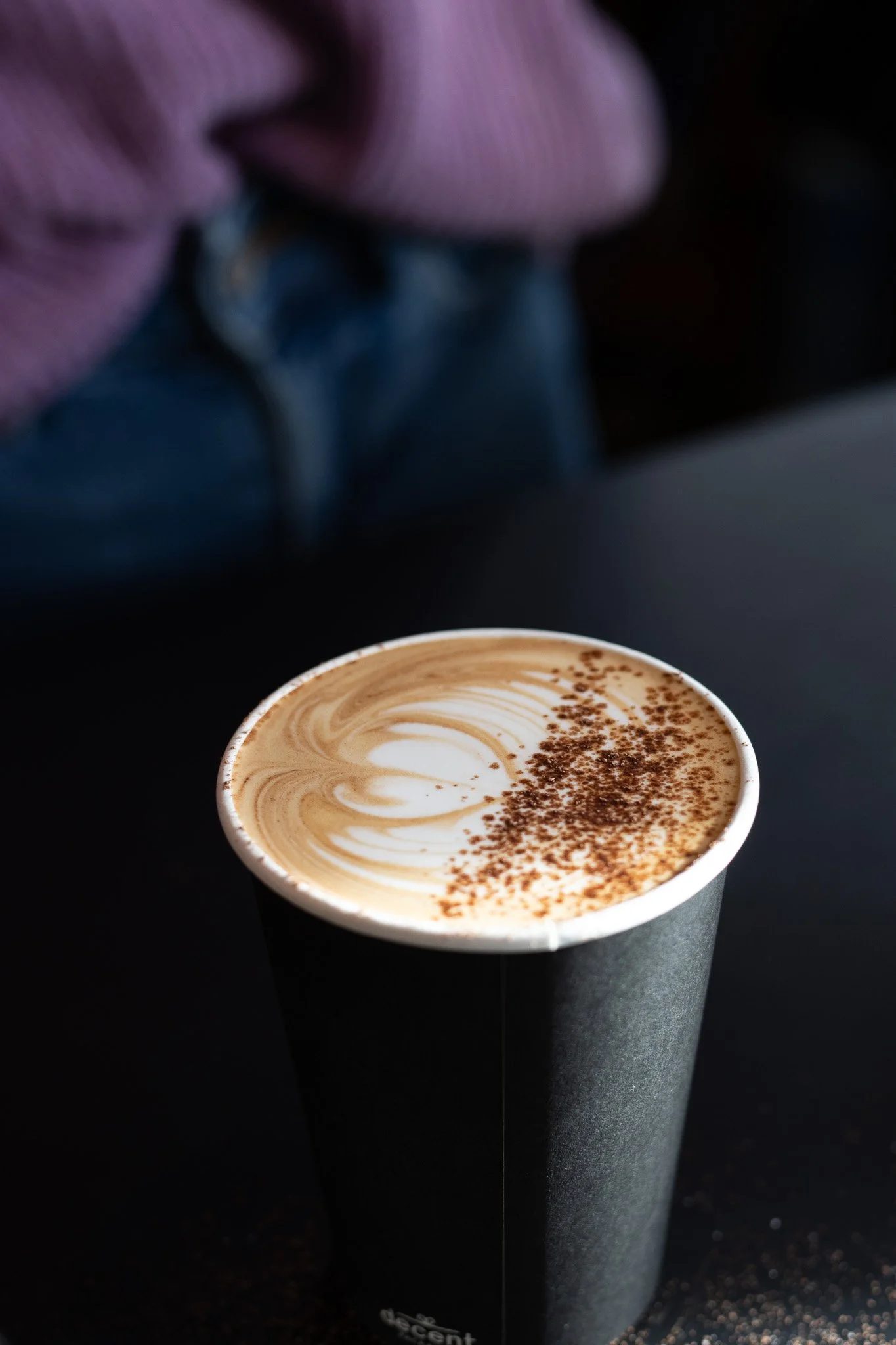 A paper cup filled with a latte, topped with swirled foam and sprinkled with cocoa powder, placed in front of a person's face. The Scandinavian Coffee Pod.
