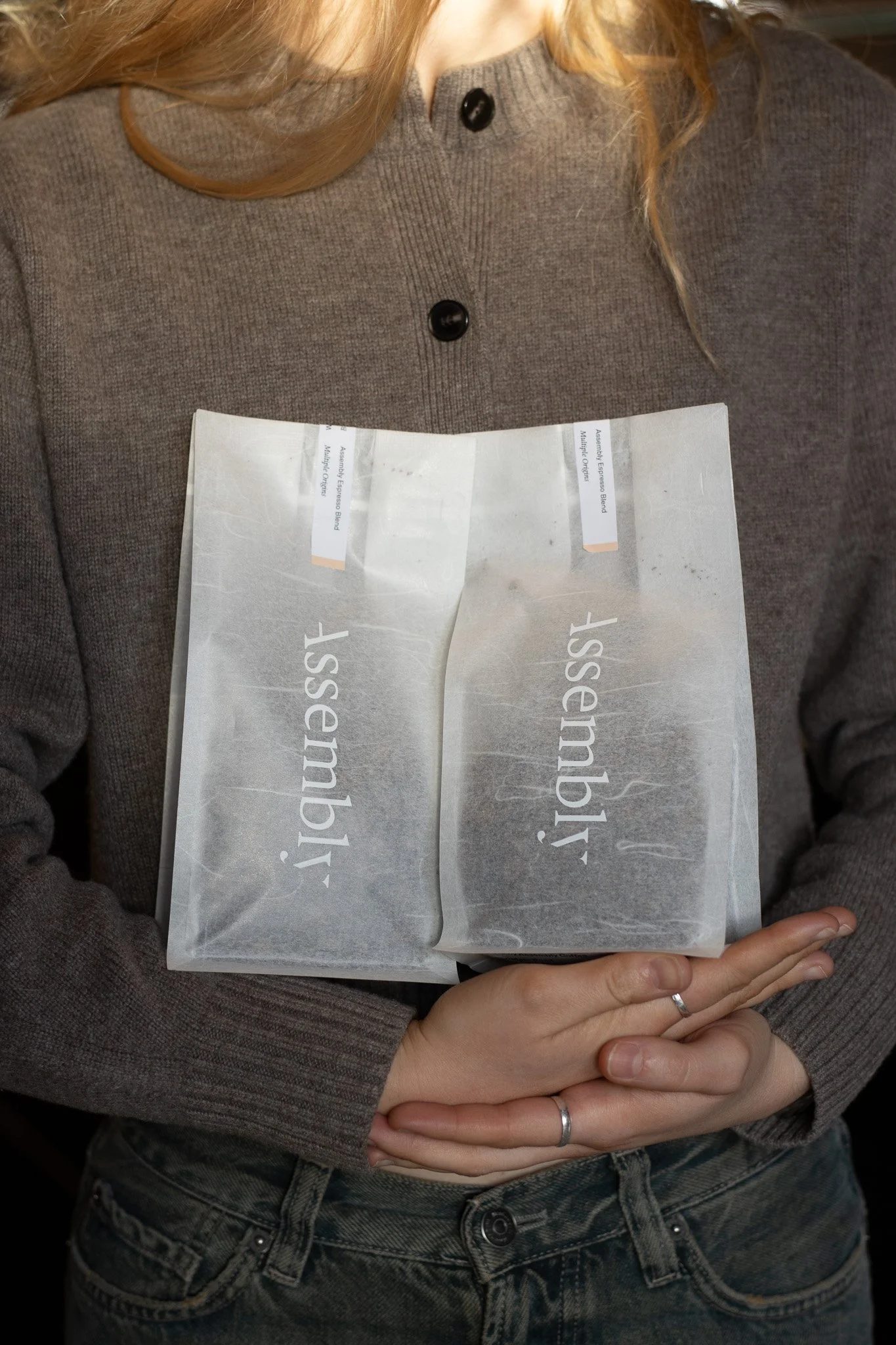 A person wearing a gray sweater and jeans is holding a plastic package labeled 'Assist' in front of their chest. The package appears to contain a product and the person has their hands clasped around it. The Scandinavian Coffee Pod.