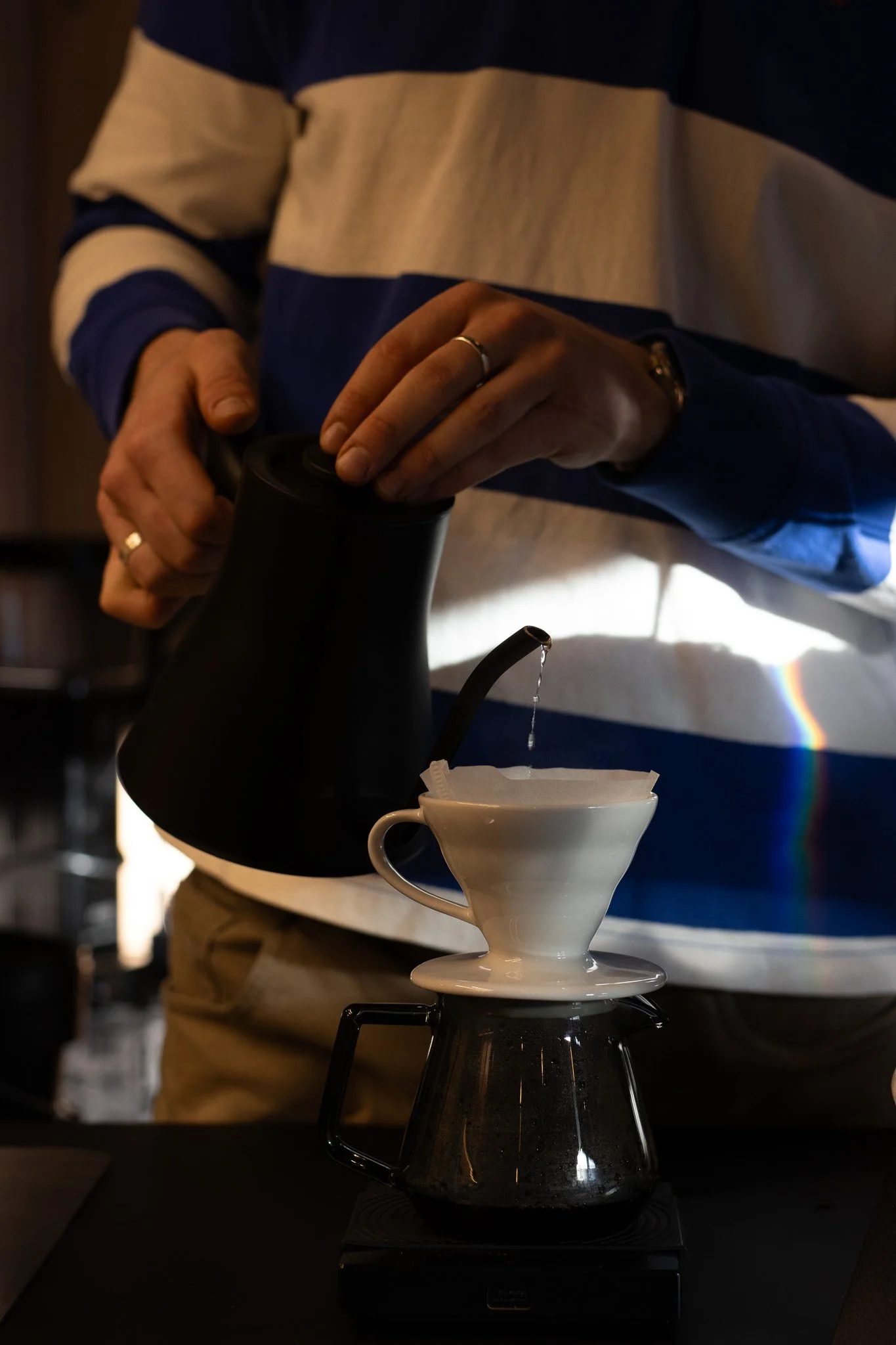 Person pouring hot water from a kettle into a white teacup on a black stove with a colorful rainbow refraction visible on the person's shirt. The Scandinavian Coffee Pod.