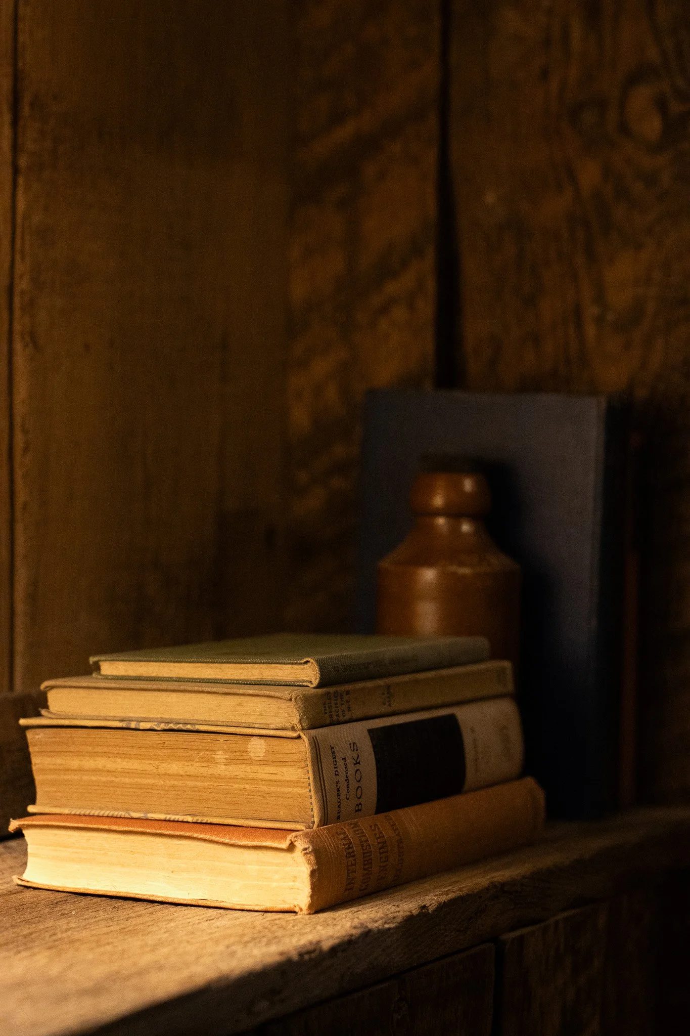 Stack of old, worn books on a wooden surface with a dark background, a small bookend and a decorative vase in the background. The Lamb Inn.