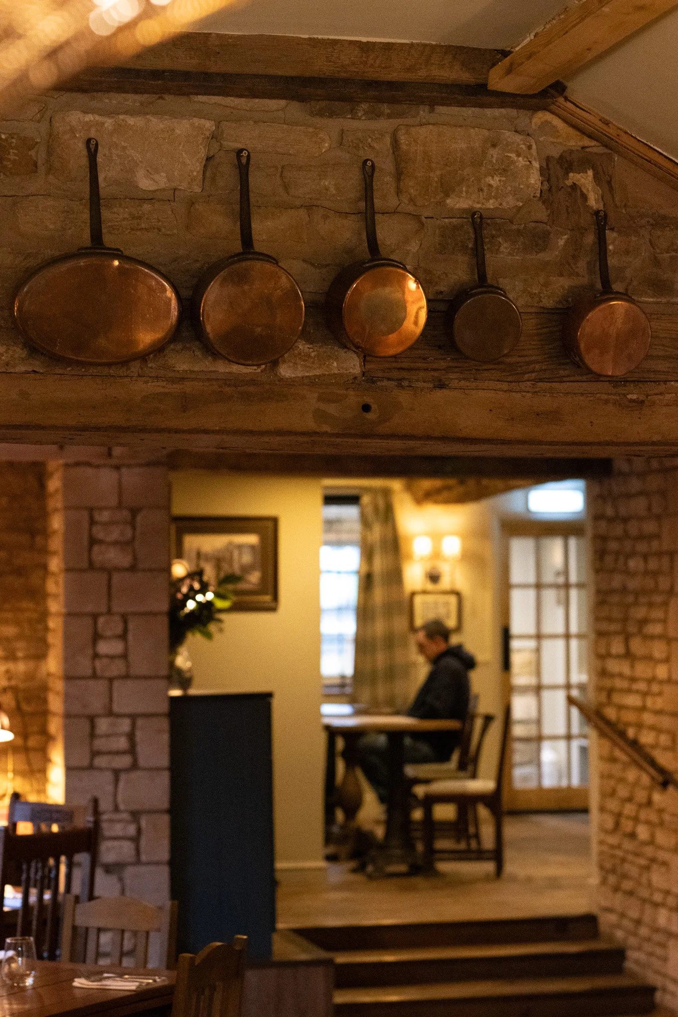 Copper pans hanging on a brick wall in a cozy restaurant or cafe, with a person sitting at a table in the background. The Lamb Inn.