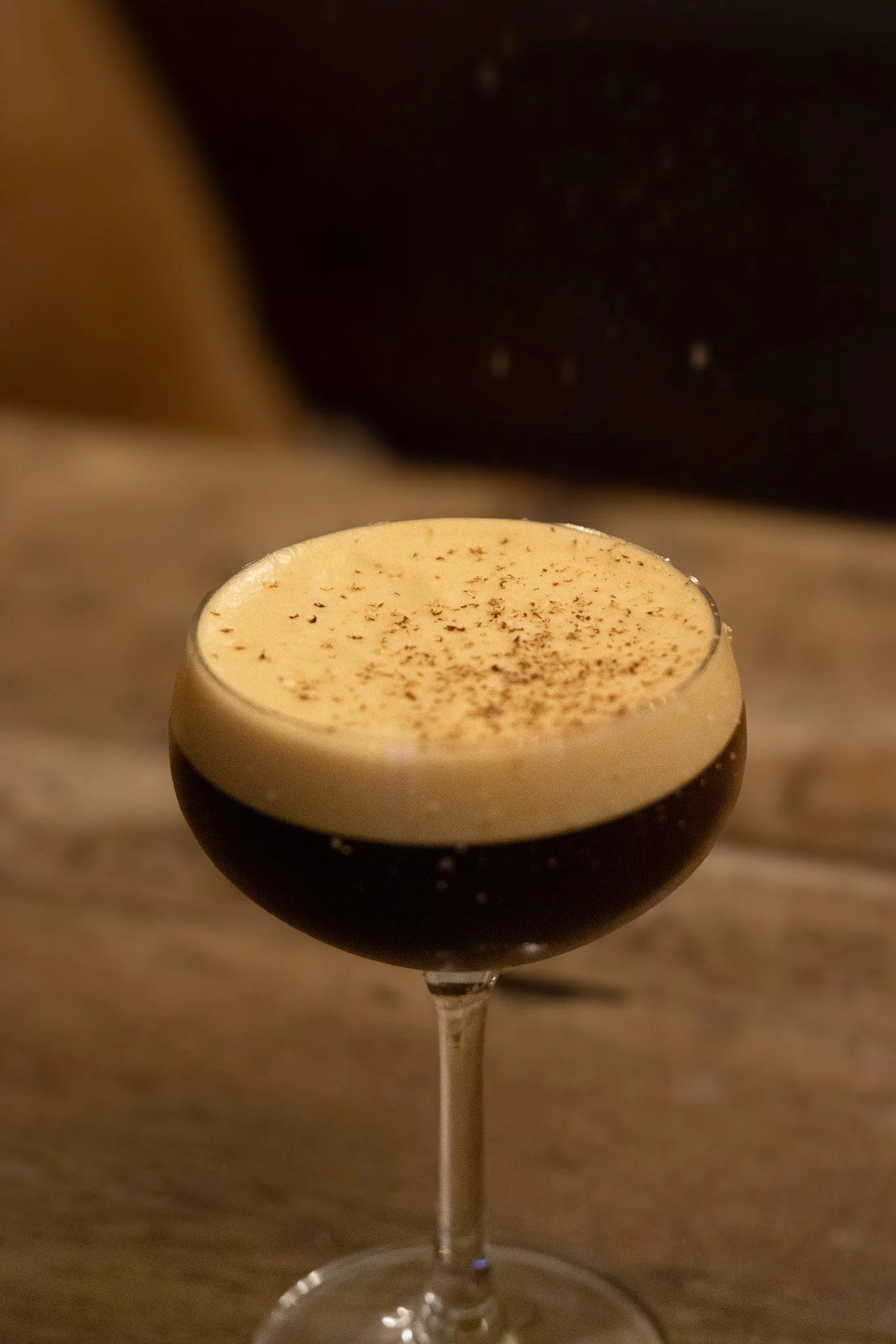 A close-up of a layered coffee drink in a cocktail glass with a dark chocolate layer at the bottom and a creamy top sprinkled with cocoa powder. The Lamb Inn. Espresso Martini.