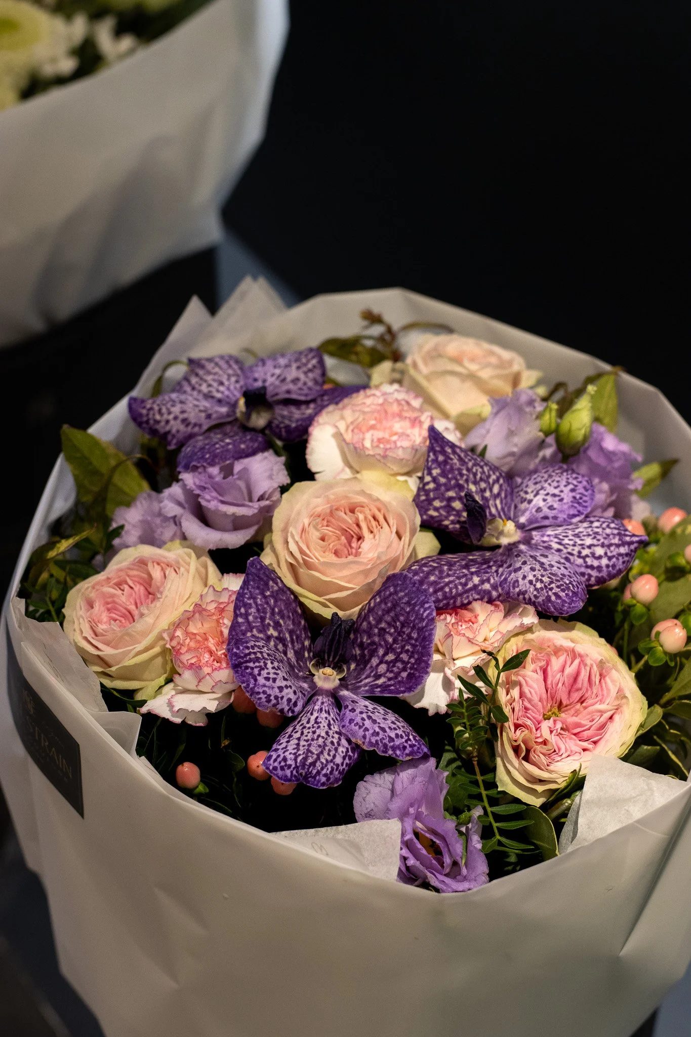 A bouquet of pink roses, purple orchids, and other purple flowers in a white wrapping paper. Neill Strain, Harrods, Motcomb Street.