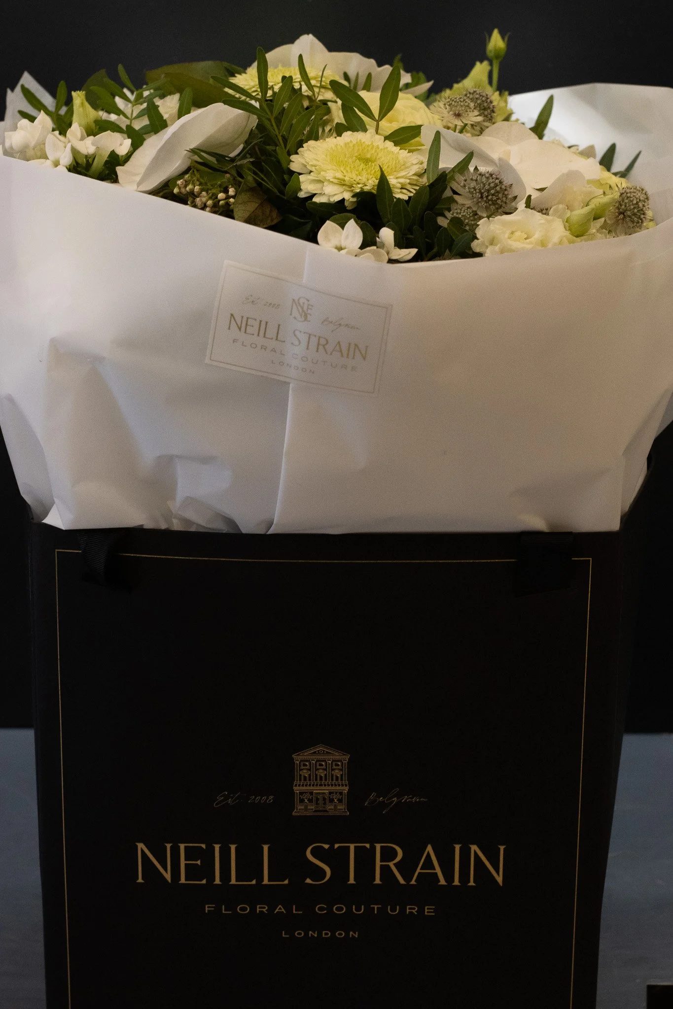 A bouquet of white flowers with greenery wrapped in white paper, labeled with a tag from Neil Strain Floral Couture London, placed on top of a black box with gold details. Neill Strain, Harrods, Motcomb Street.