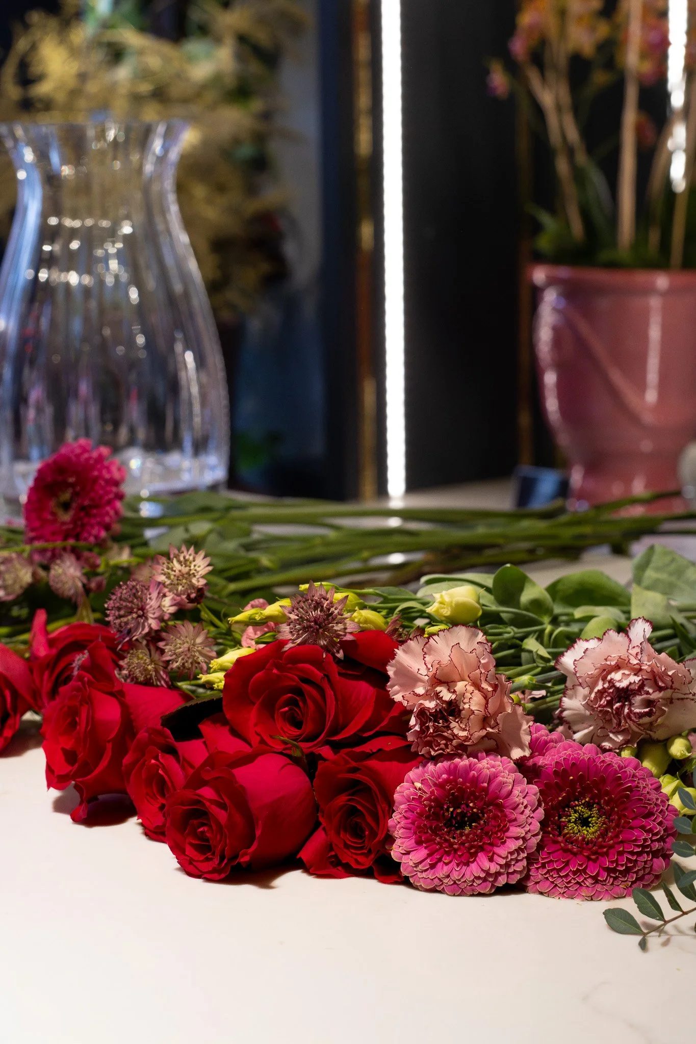 A variety of colorful flowers, including roses and carnations, arranged on a white surface with a blurred background of glass vases and potted plants. Neill Strain, Harrods, Motcomb Street.