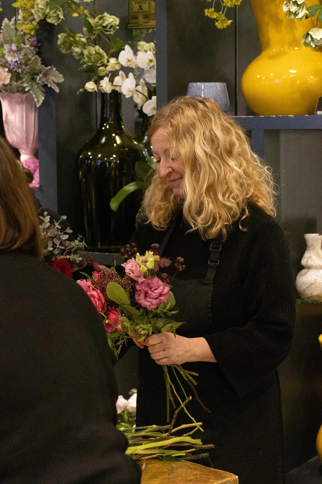 A woman with curly blonde hair, wearing a black top, is holding a bouquet of pink, purple, and green flowers. She is smiling softly and looking down at the flowers. Behind her, there are large decorative vases with white and yellow flowers, and a sec