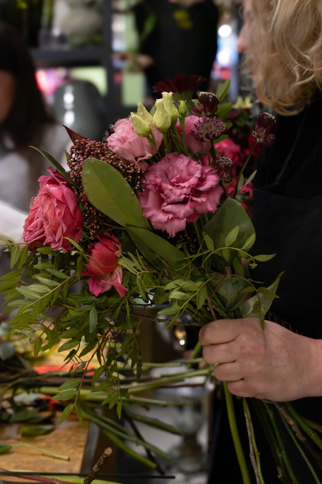 A person with curly blonde hair arranging pink and purple flowers with green leaves at a market or floral shop. Neill Strain, Harrods, Motcomb Street.