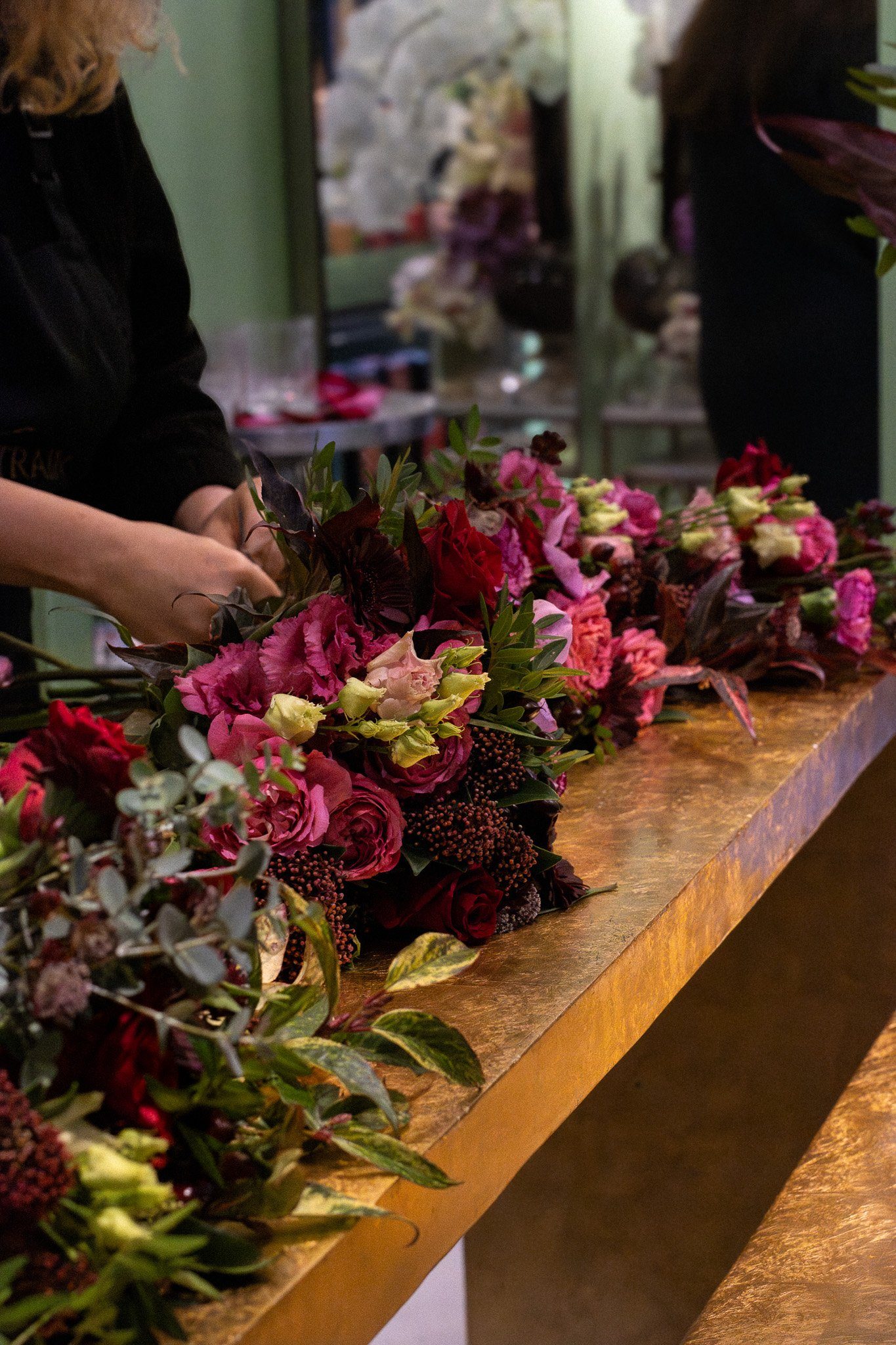 A person arranging a variety of colorful flowers on a wooden table in an indoor setting, with other people in the background. Neill Strain, Harrods, Motcomb Street.