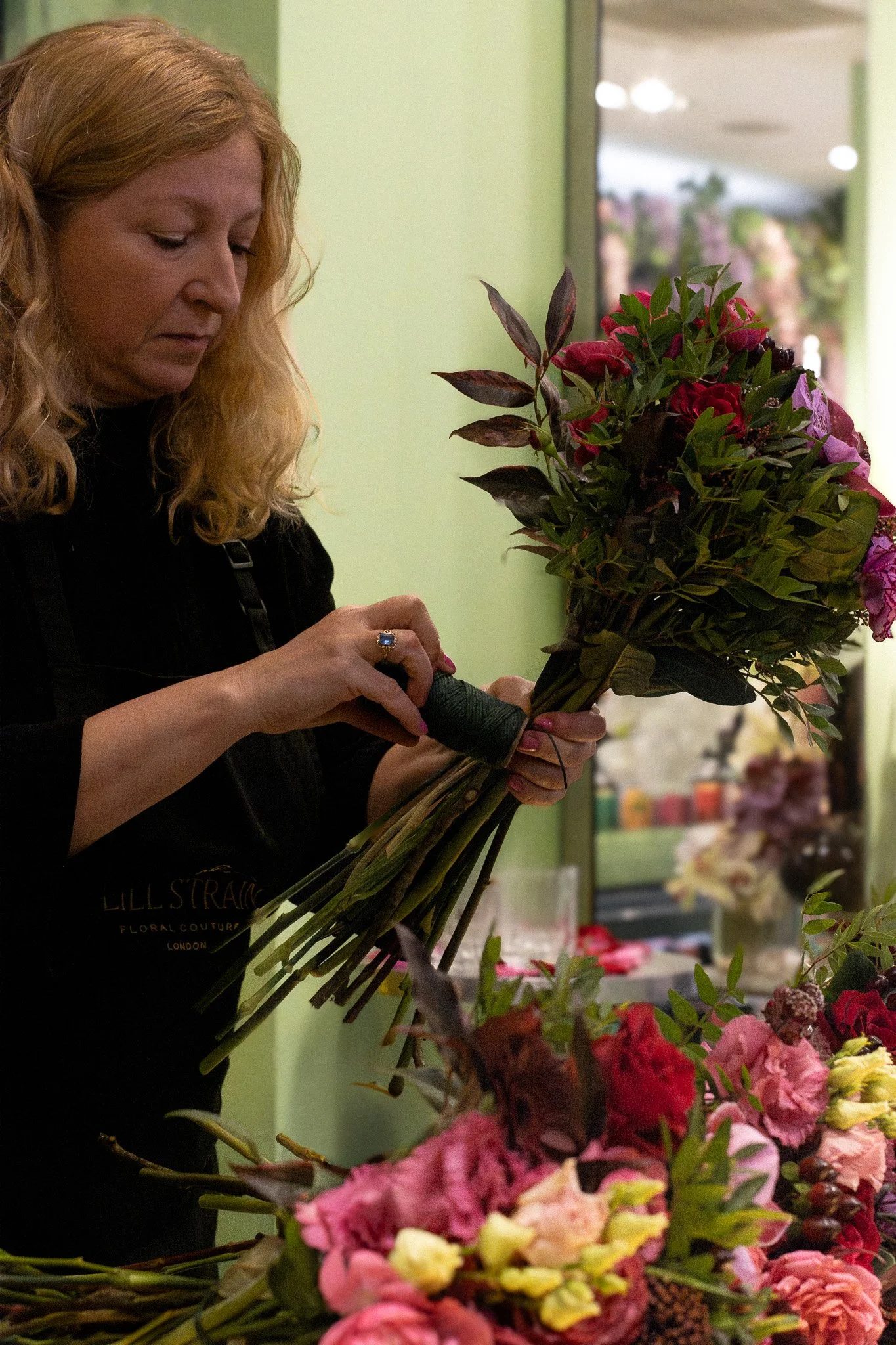 A woman with long blonde hair arranging a large bouquet of mixed flowers, including roses, in a flower shop with green walls and blurred background. Neill Strain, Harrods, Motcomb Street.