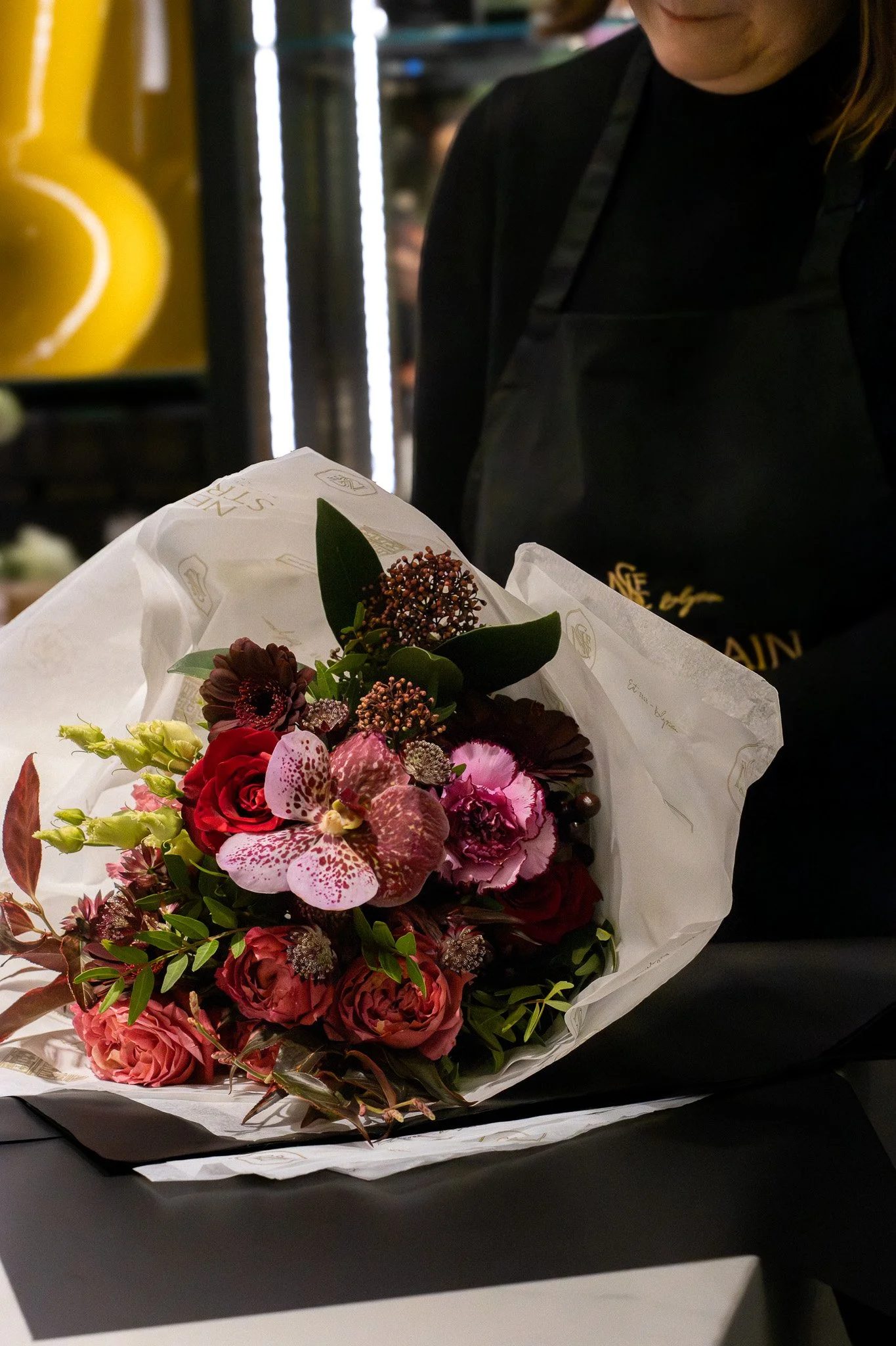 A woman in black clothing holding a colorful flower bouquet wrapped in white paper. Neill Strain, Harrods, Motcomb Street.