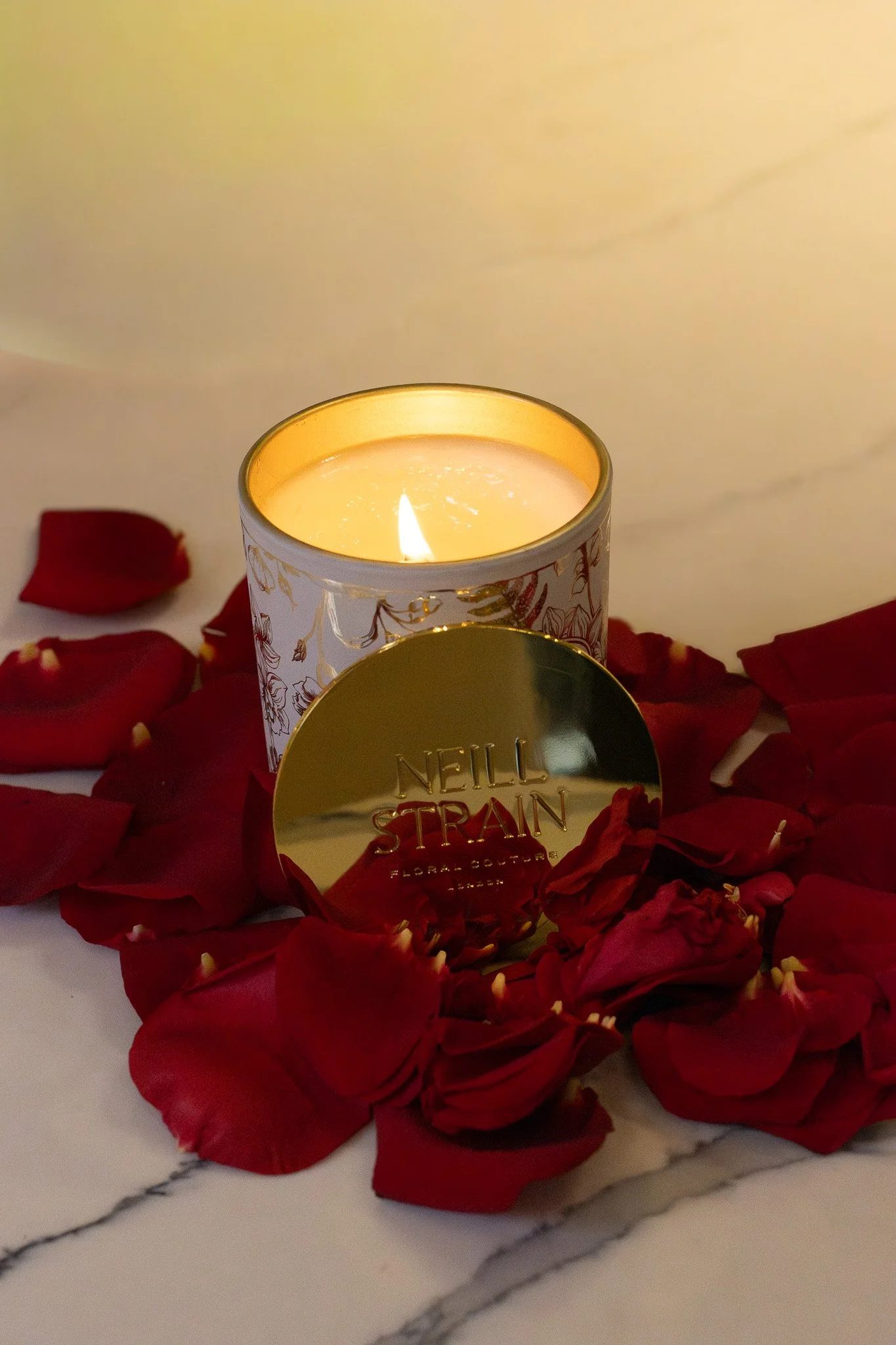 A lit candle in a decorative container surrounded by red rose petals, with a gold foil box labeled 'Neil Grain Floral Bouquets' partially visible in the foreground, on a marble surface. Neill Strain, Harrods, Motcomb Street.
