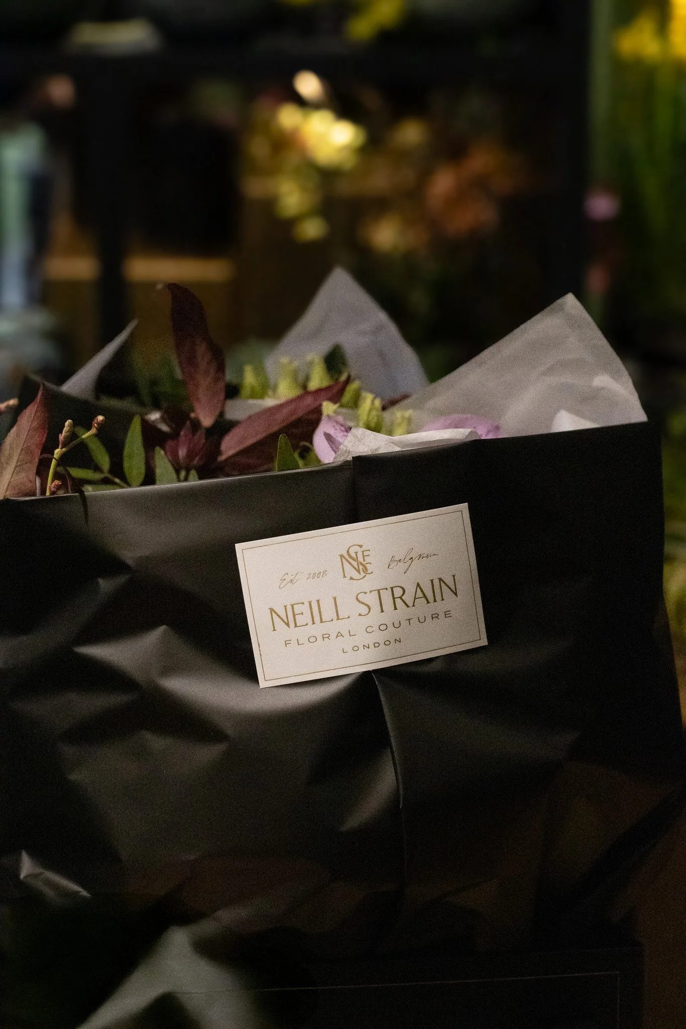 A black gift bag with a white label from Neill Strain Floral Couture in London, containing flowers and foliage, with a blurred background. Neill Strain, Harrods, Motcomb Street.