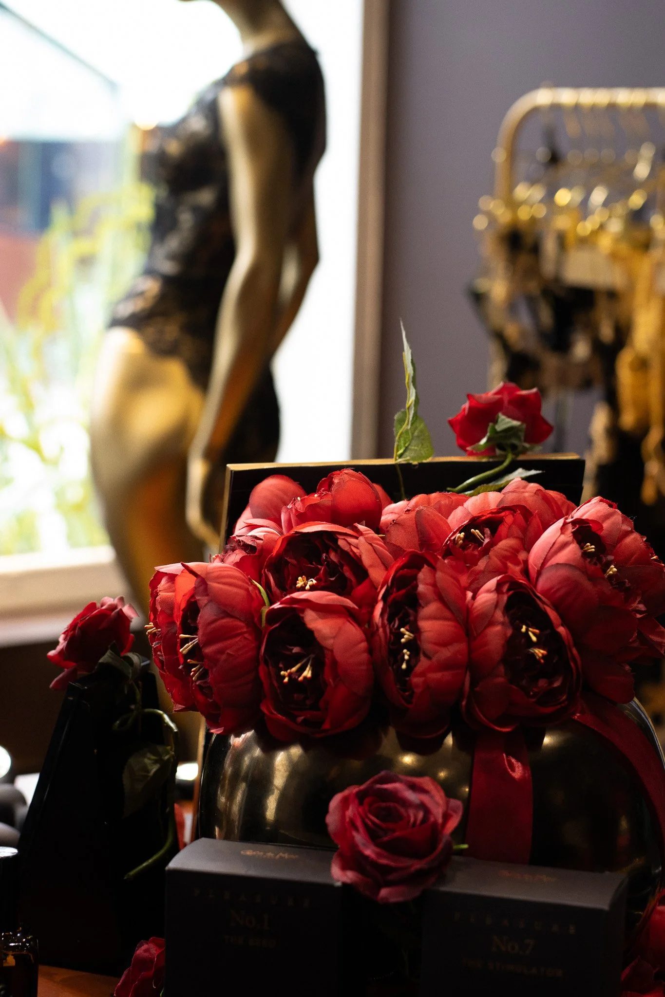 A close-up of a vibrant red rose bouquet in a dark metallic vase in front of a black box with gold text, with a gold decorative piece and blurry artwork, with a window to the left providing natural light. Coco De Mer.
