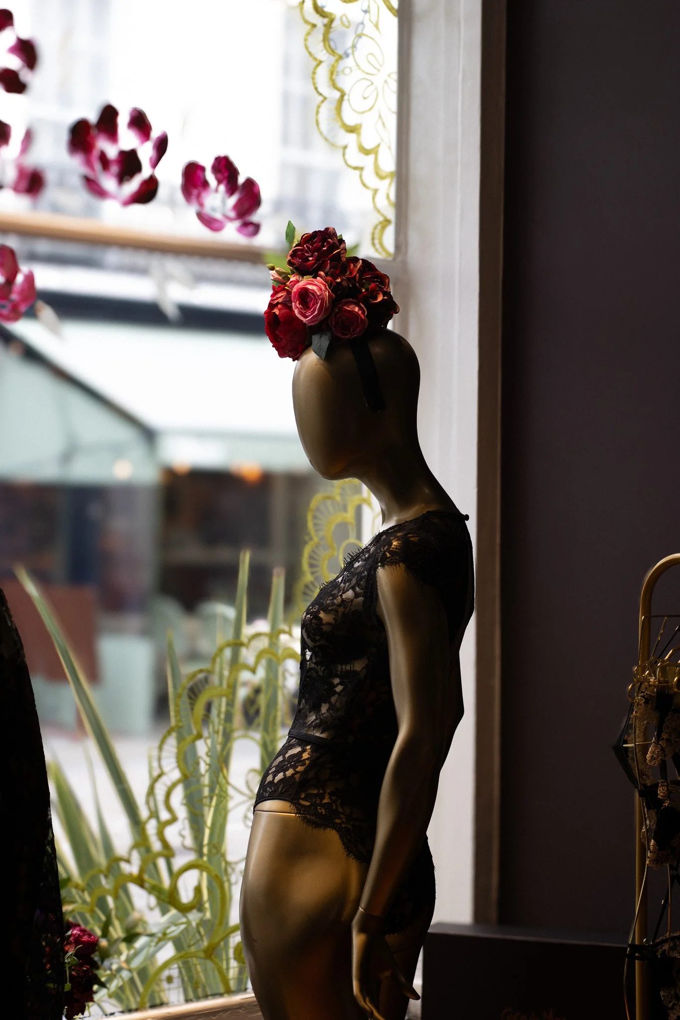 A mannequin dressed in black lace lingerie, with a floral headpiece, standing indoors near a window with lace curtains, surrounded by plants. Coco De Mer.