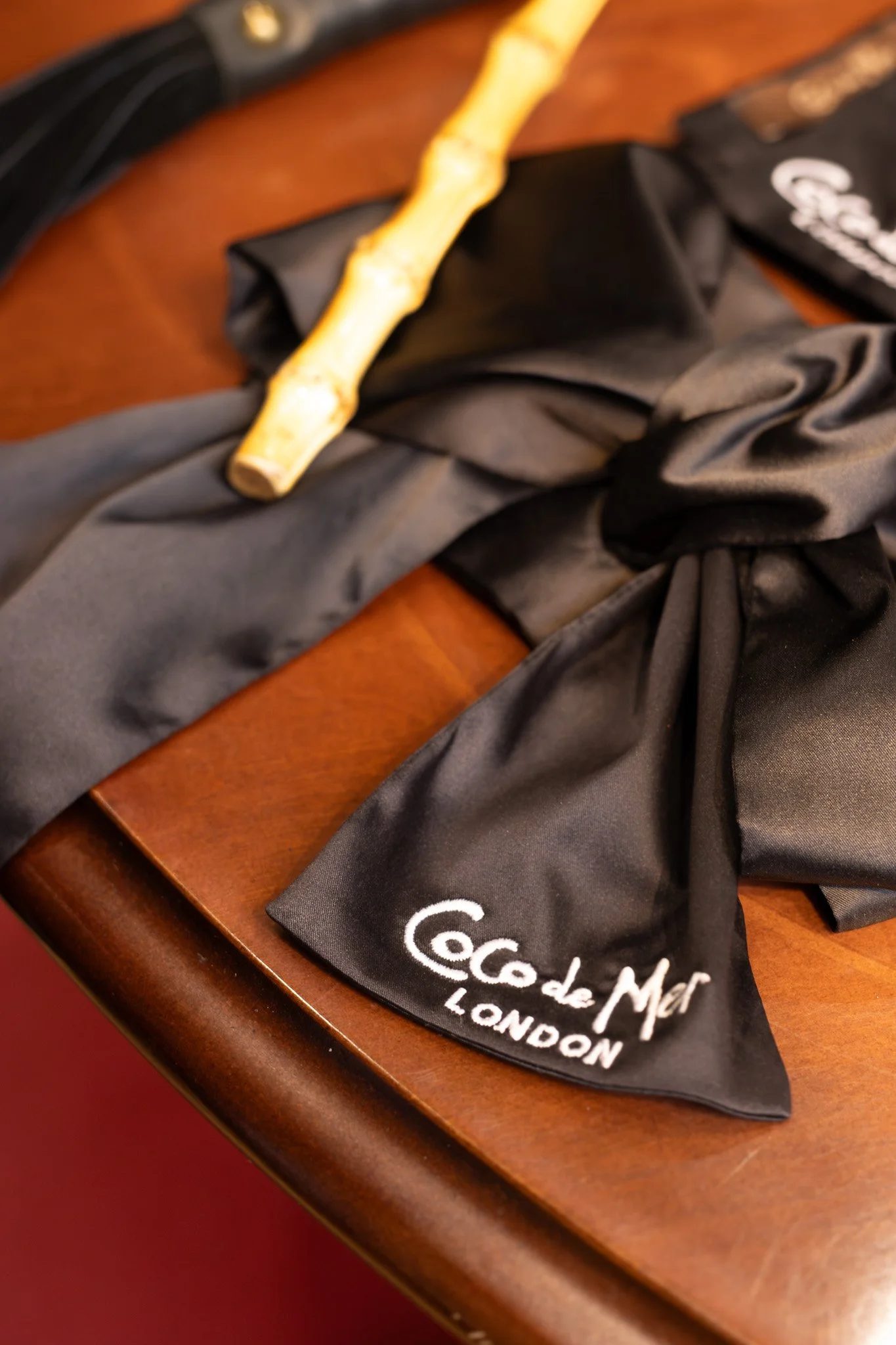 Black silk scarf with white embroidery reading "Coc de Mar LONDON" on a wooden surface, with a bamboo stick and some black wired object nearby. Coco De Mer.