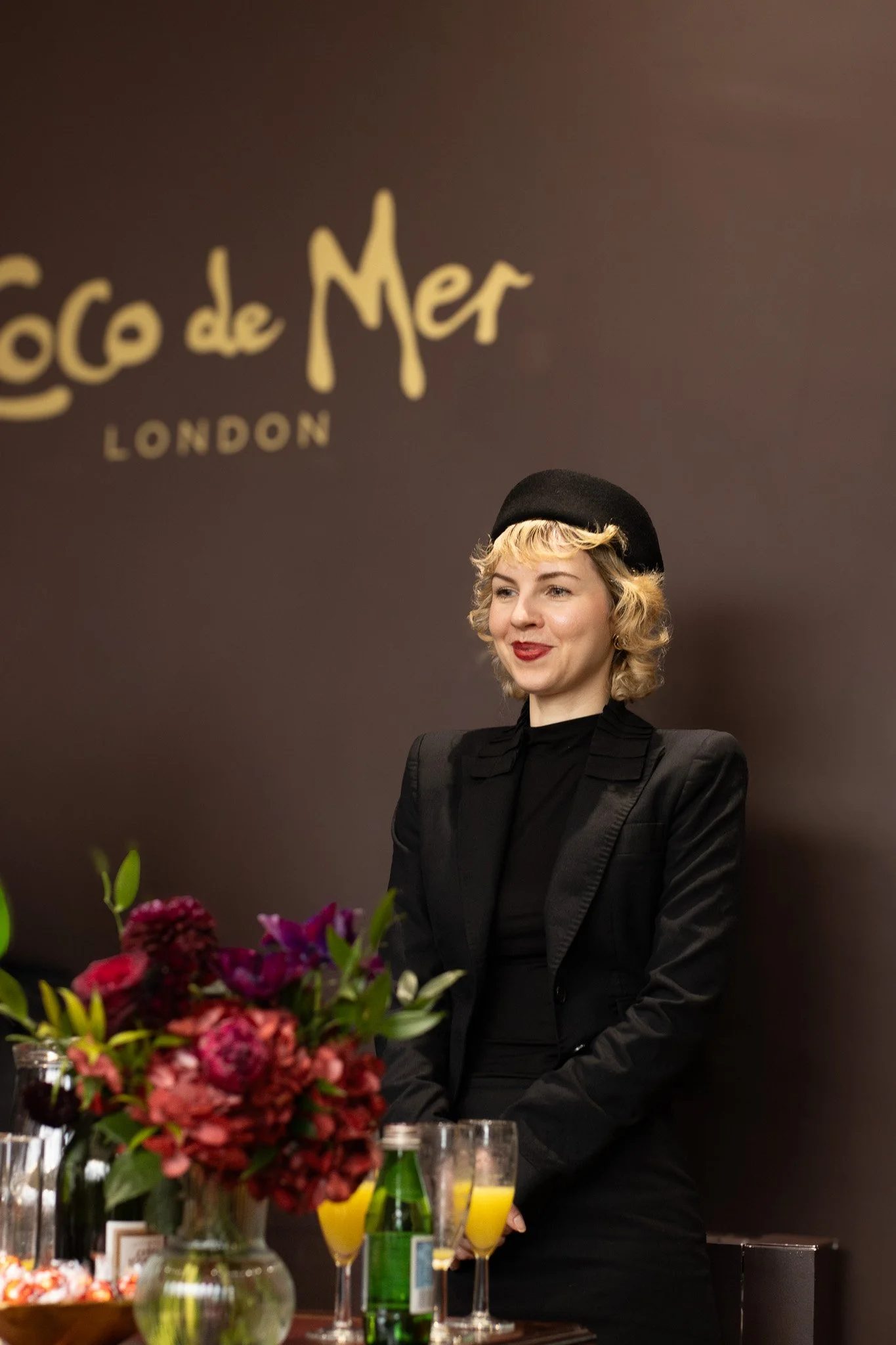 A woman with blonde curly hair and red lipstick wearing a black beret and black blazer standing behind a table with a bouquet of flowers, glasses of orange juice, and bottles, in front of a brown wall with gold text that reads 'Coco de Mer London.'