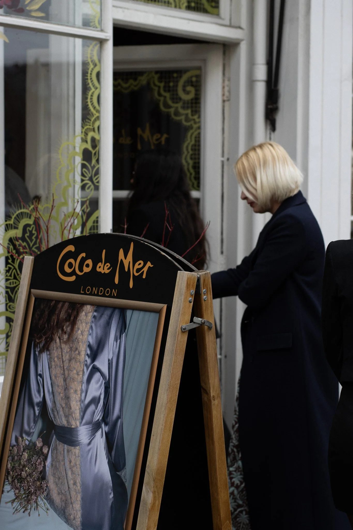 A woman with blonde hair wearing a black jacket is standing outside a shop window, looking down. Another person with dark hair is inside the shop, facing away. A sign in front of the shop reads: "Coco de Mer London." The window displays decorative ye
