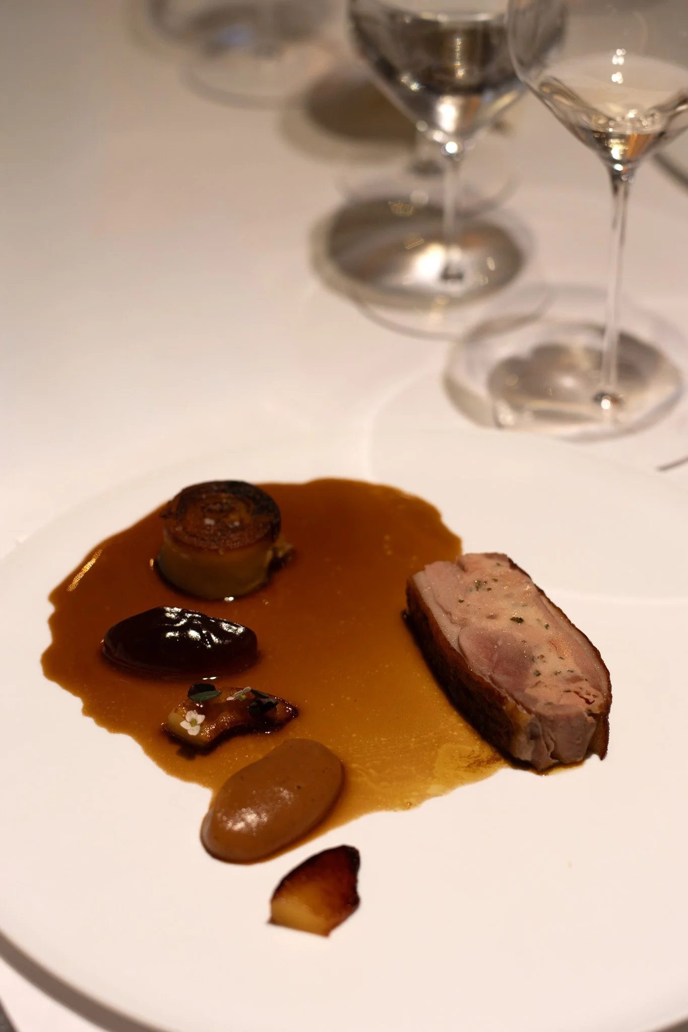 Close-up of gourmet appetizer featuring sliced meat, sauces, and garnishes on a white plate, with empty wine glasses in the background. Petrus.