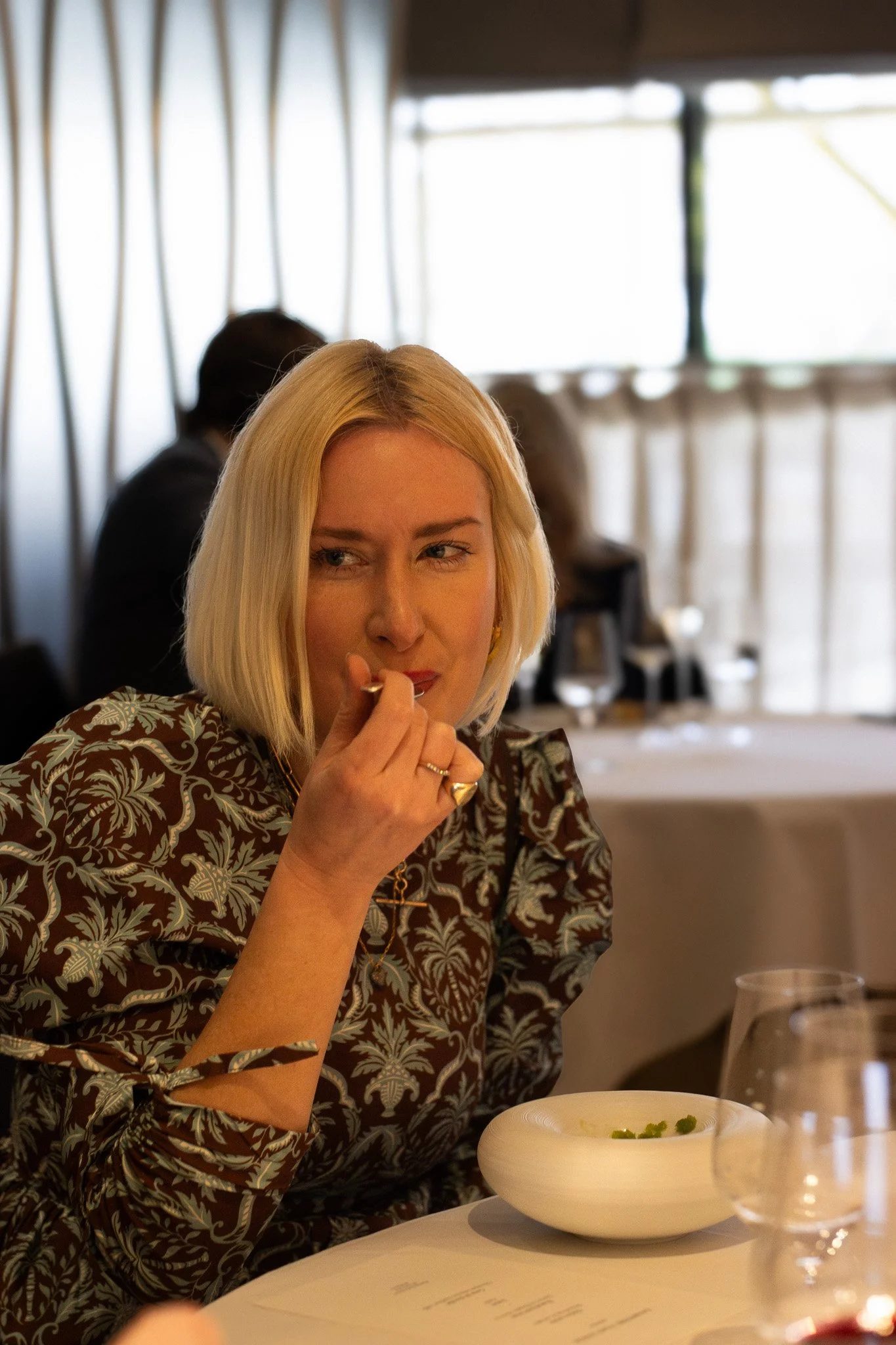 A woman with blonde hair sitting at a dining table, looking to her left, with a thoughtful expression. In front of her is a white bowl of soup garnished with herbs, and there are wine glasses on the table. Petrus.