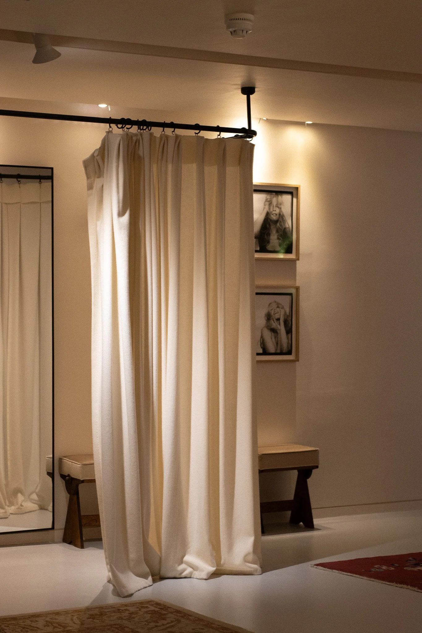A room with white curtains hanging from a black curtain rod, a framed black-and-white portrait of a woman with long hair and a hand on her head, a small wooden side table, and a patterned area rug on the floor. Realisation Par.
