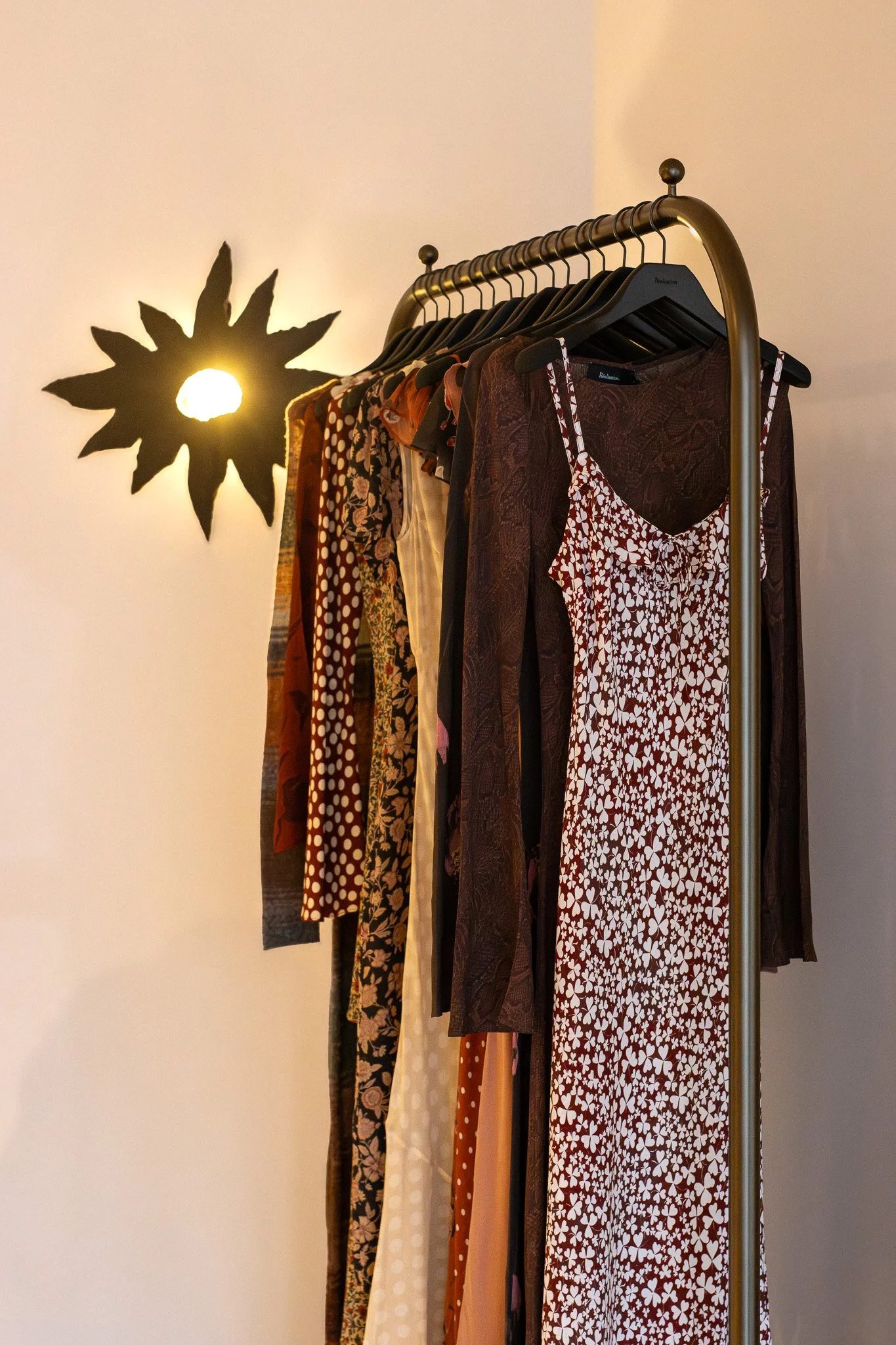 Clothing rack with various dresses and garments hanging, illuminated by a wall-mounted light fixture shaped like a sunflower. Realisation Par.
