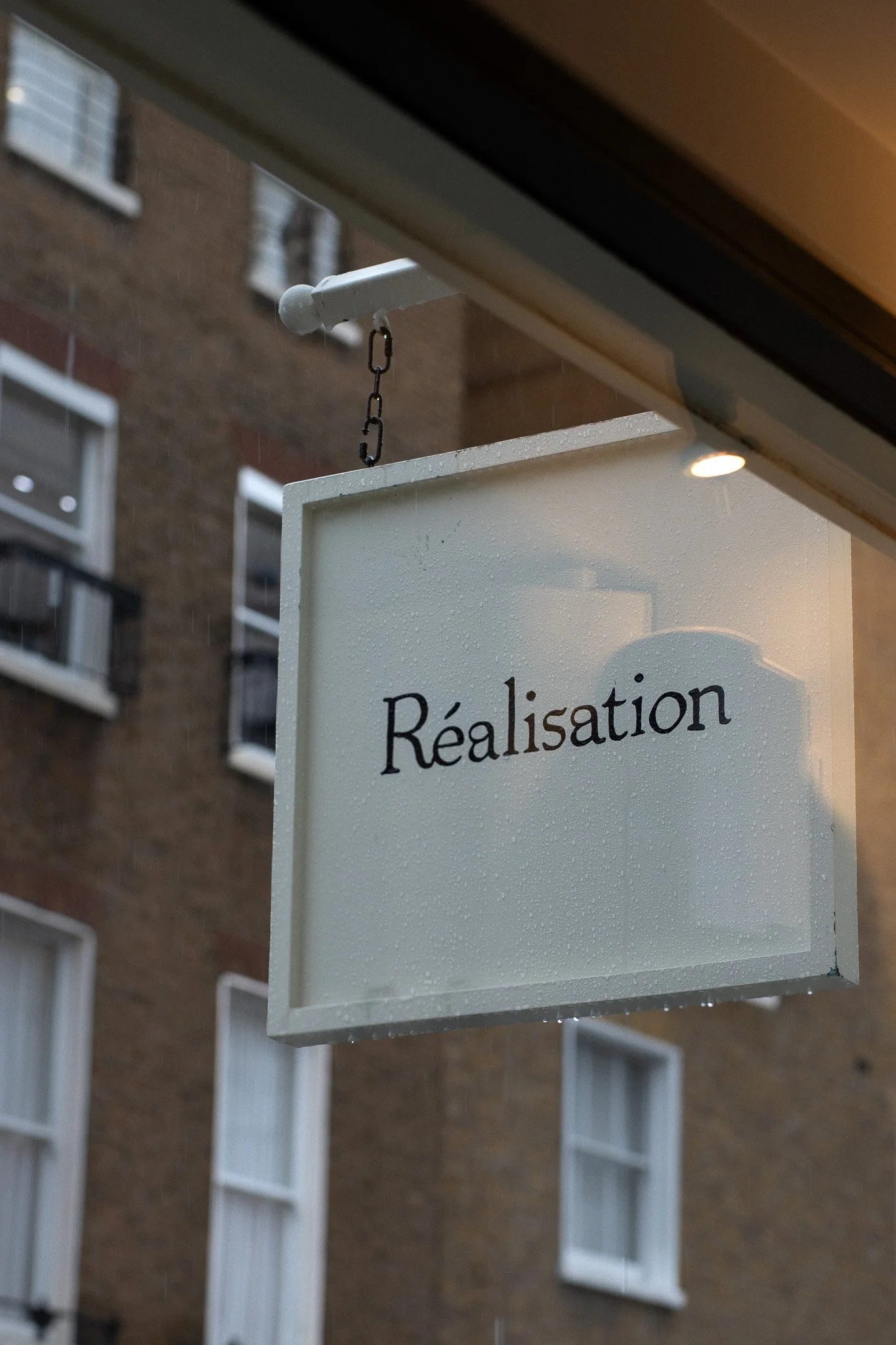 A white sign with black text that reads 'Réalisation,' hanging outside a building. The sign is wet with rain and attached to a chain and metal arm extending from the building's overhang. Realisation Par.