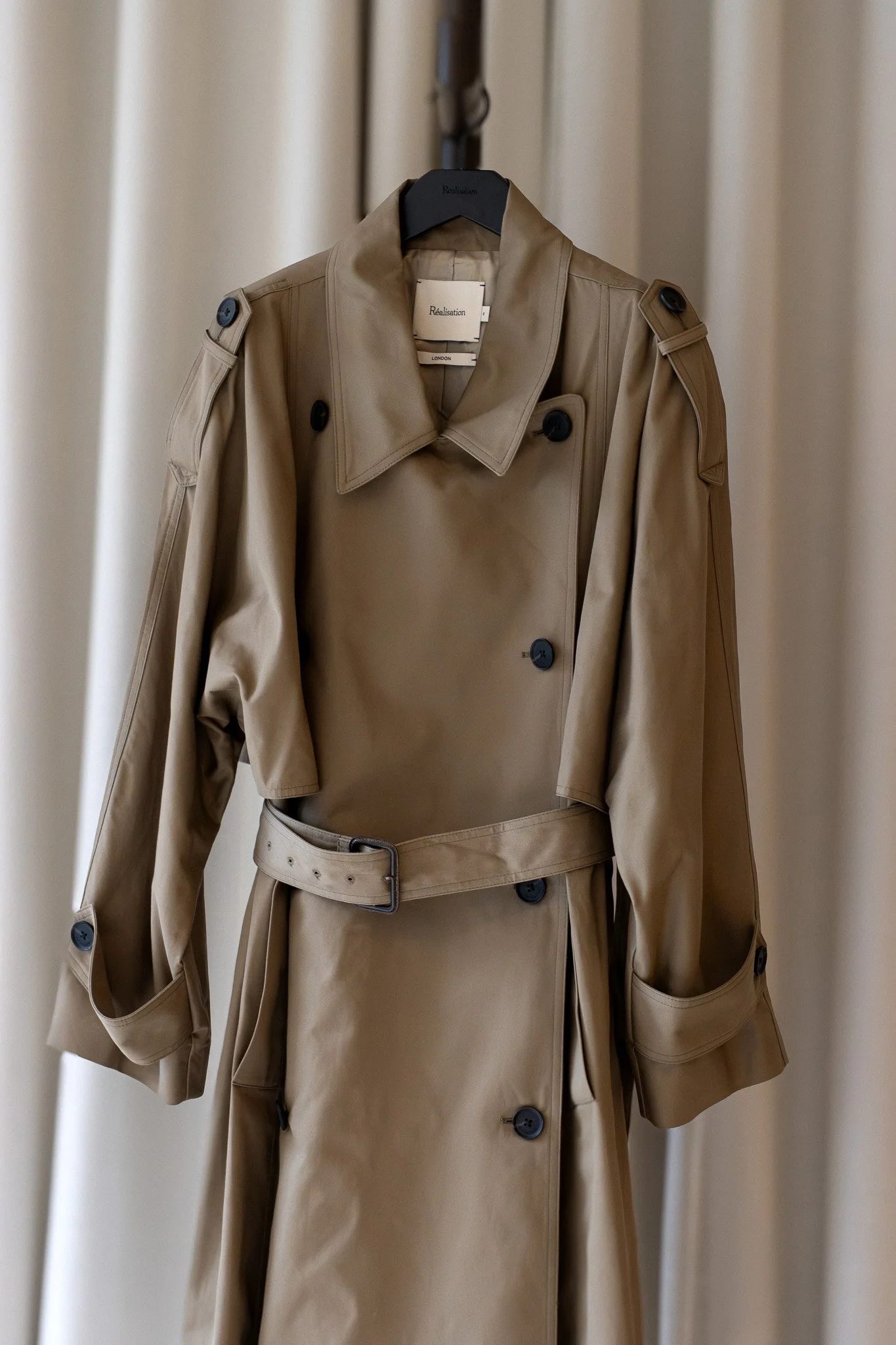 A beige trench coat hanging on a black hanger in front of a light-colored curtain. Realisation Par.
