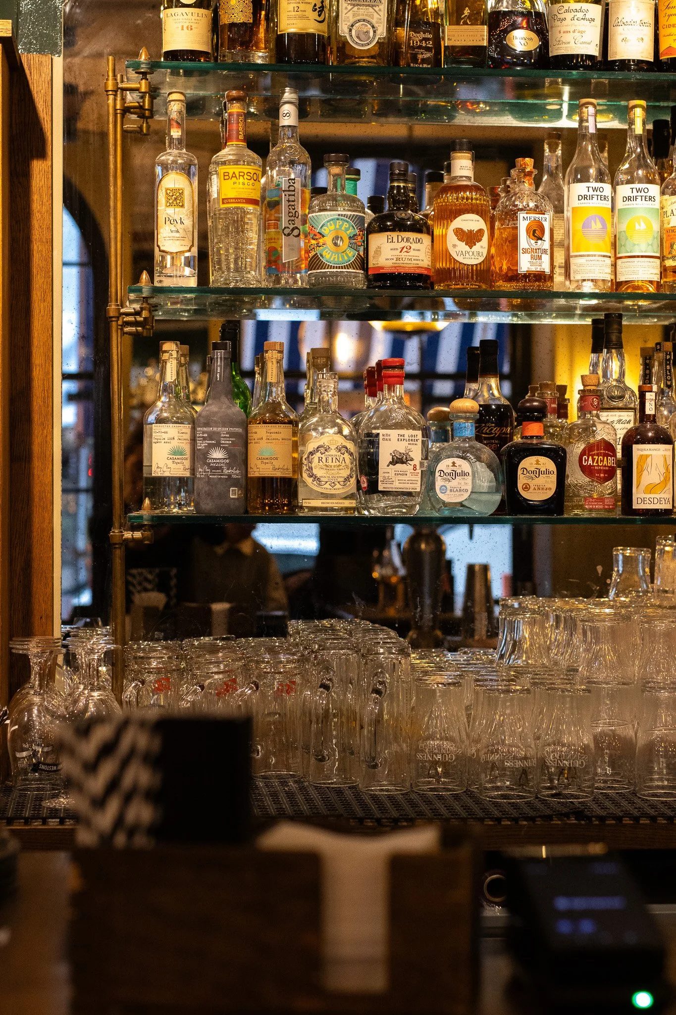 Bar shelf with various liquor bottles including whiskey, rum, and gin, along with upside-down glassware. Alfred Tennyson.