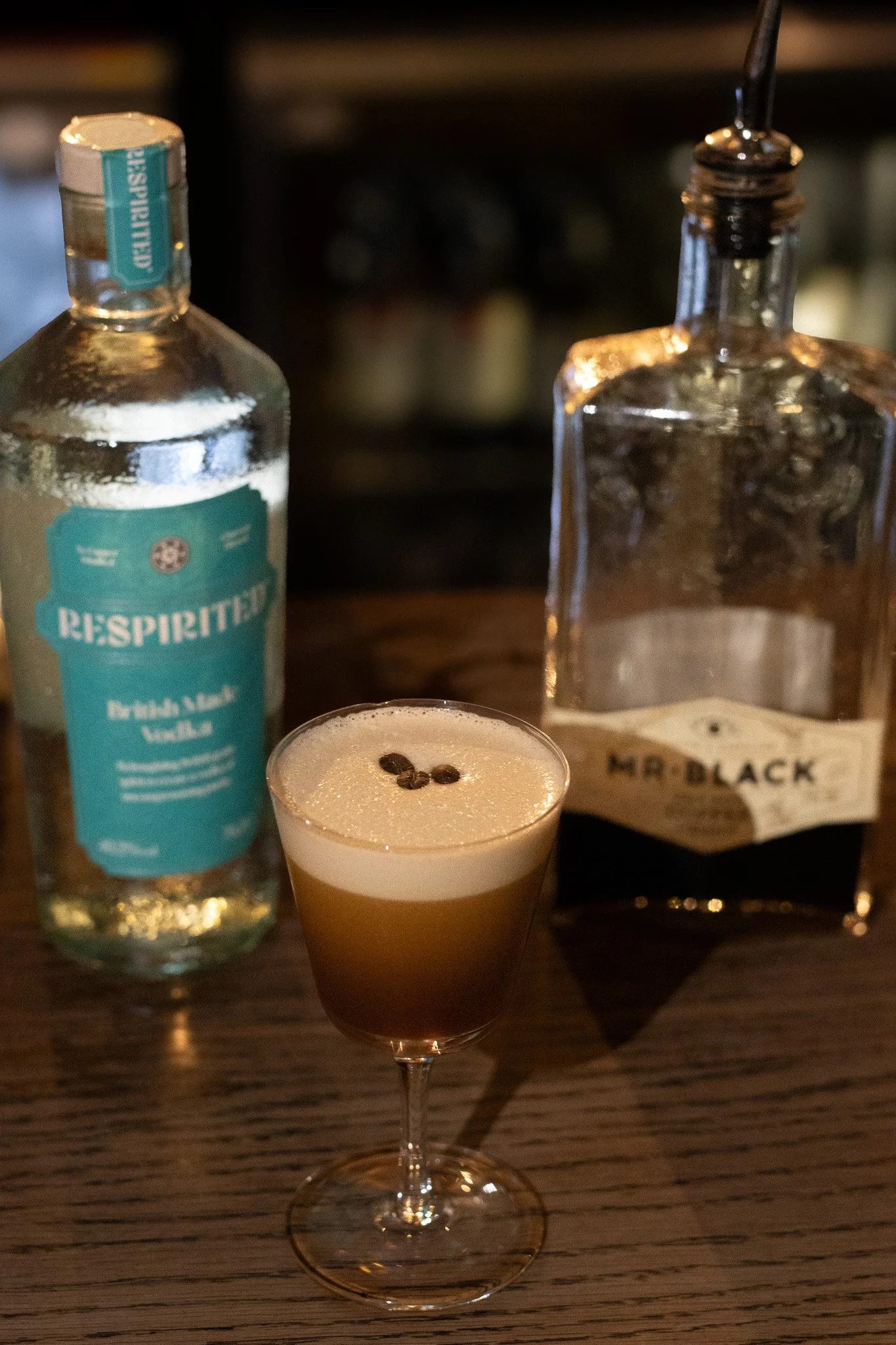 A cocktail topped with coffee beans in front of a bottle of ReSpirit and a bottle of Mr. Black Nero Coffee Liqueur on a wooden table. Alfred Tennyson.