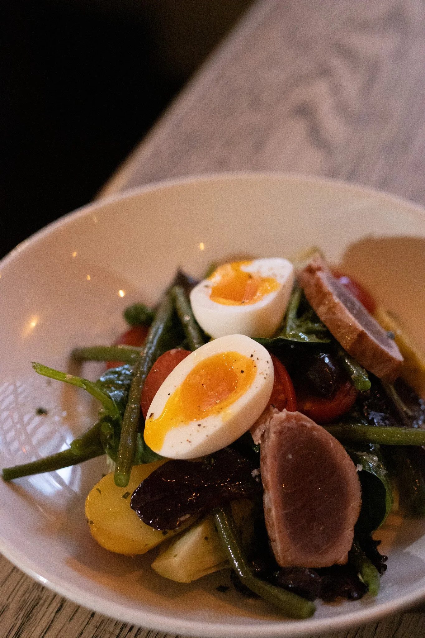 Close-up of a bowl of salad containing halved soft-boiled eggs, green beans, cherry tomatoes, slices of meat, and yellow bell peppers. Alfred Tennyson.