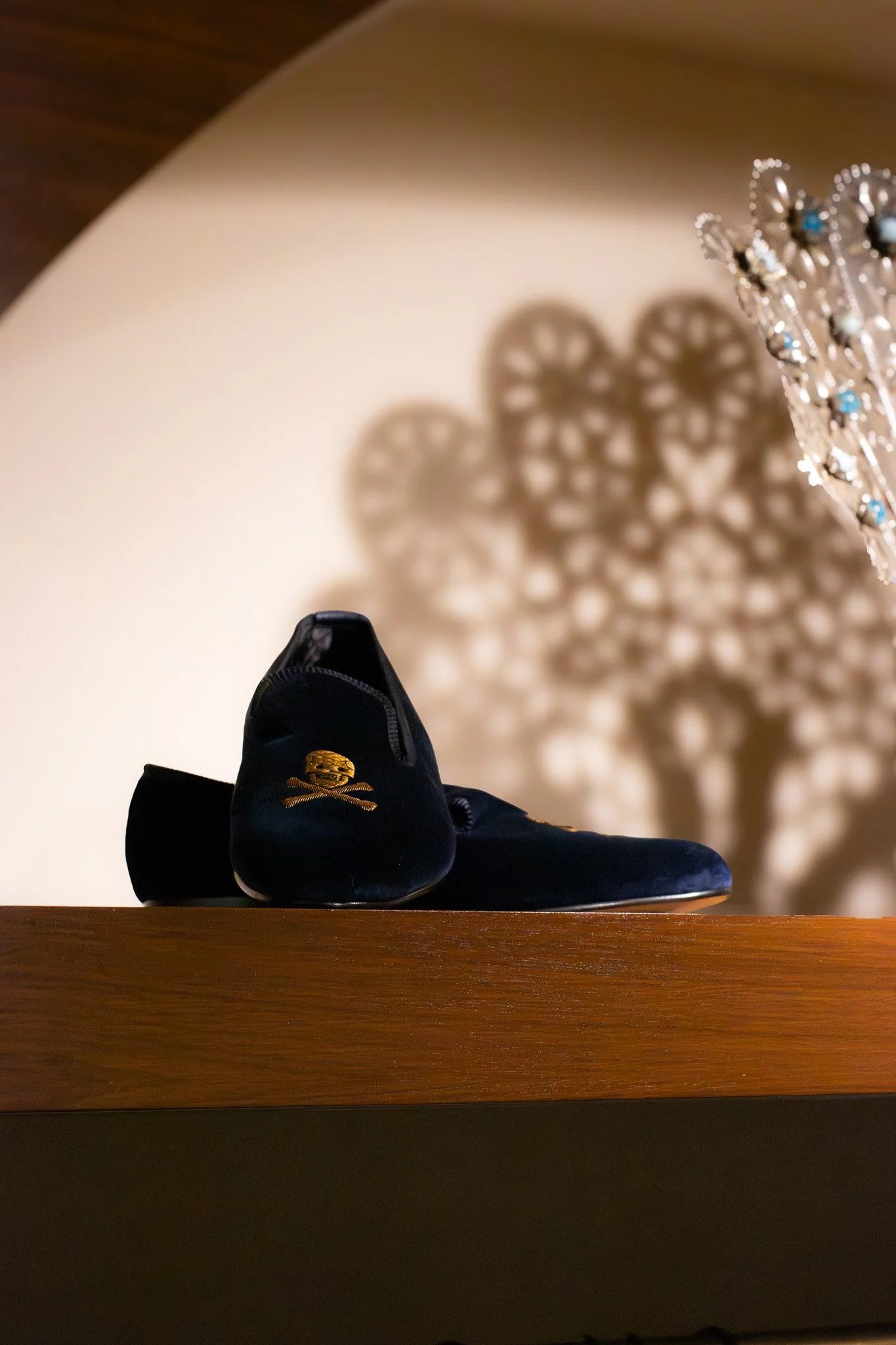 A pair of black shoes and a black hat with a gold decoration resting on a wooden surface. The shadow of a decorative object with circular patterns is cast on the wall behind them, creating a lace-like pattern. New & Lingwood.