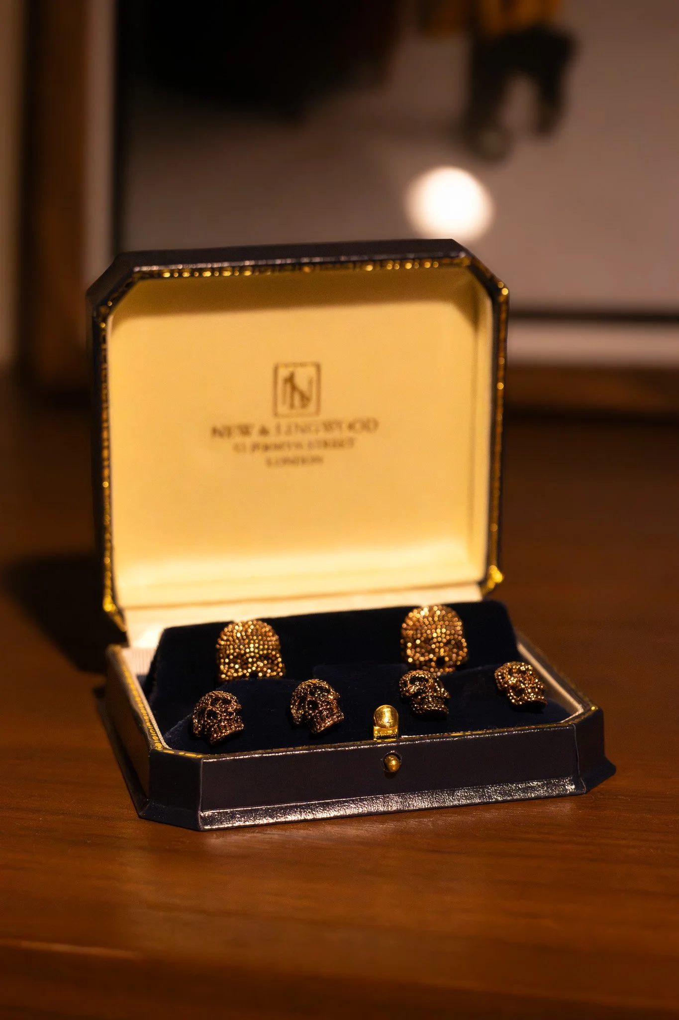 A jewelry box with gold skull-shaped earrings inside, placed on a wooden surface with a blurred background and a light source creating a warm glow. New & Lingwood.