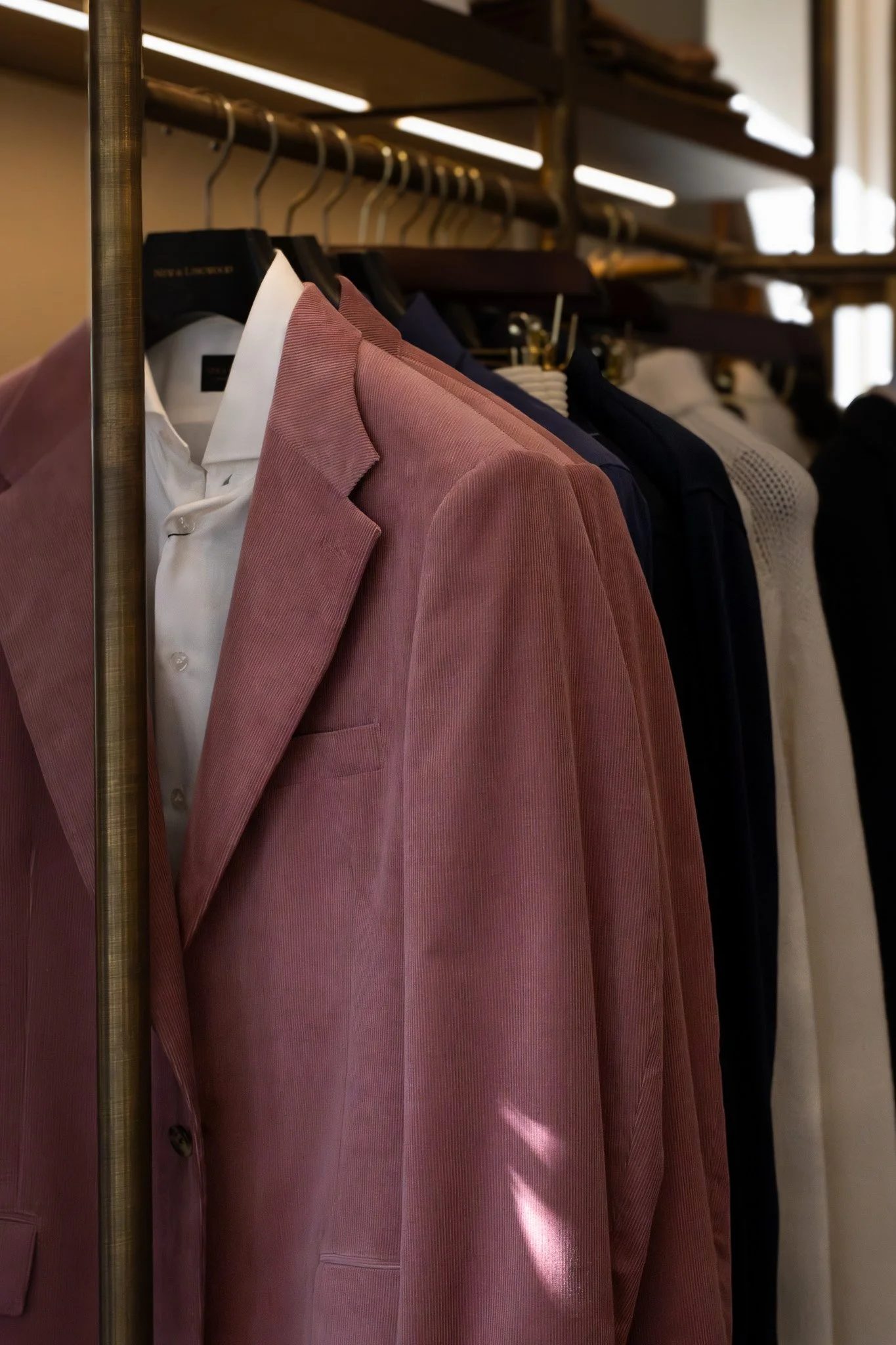 Pink blazer, white shirt, and other clothing items hanging on a clothing rack in a store or closet. New & Lingwood.