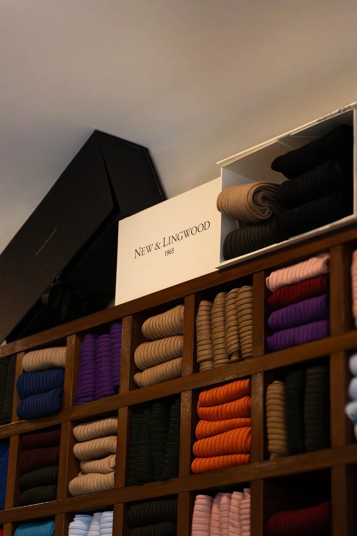 A retail display of neatly rolled striped fabrics in various colors including beige, purple, pink, orange, and black, arranged in a wooden grid shelving unit. New & Lingwood.