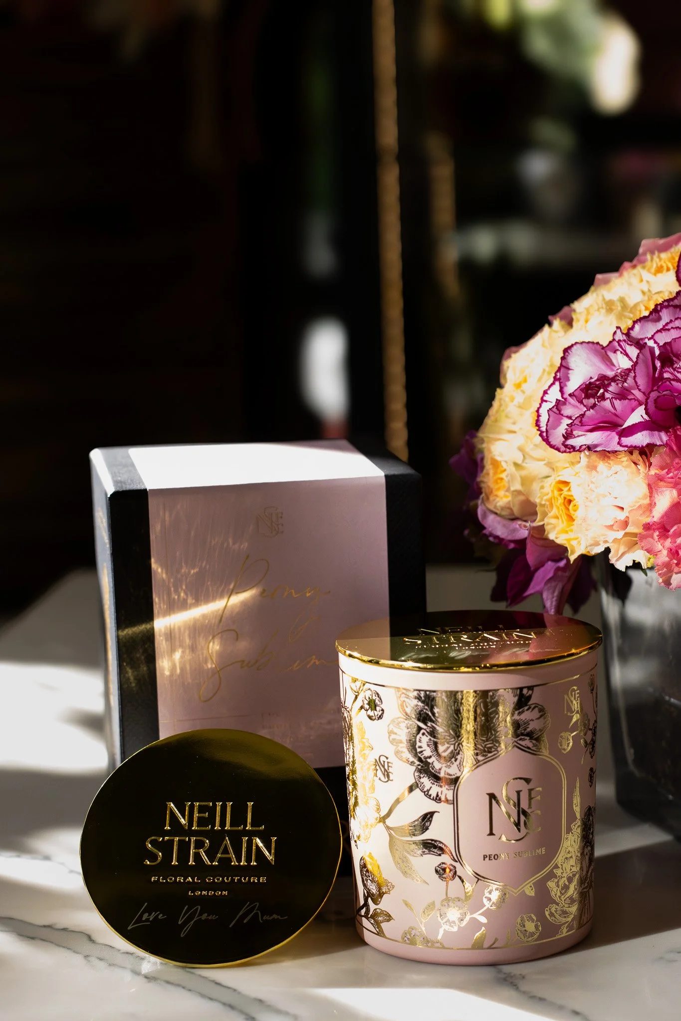 A collection of luxury products including a pink floral scented candle, a black and gold container with the name Neill Strain, and a pink box with gold accents. Neill Strain, Harrods, Motcomb Street.