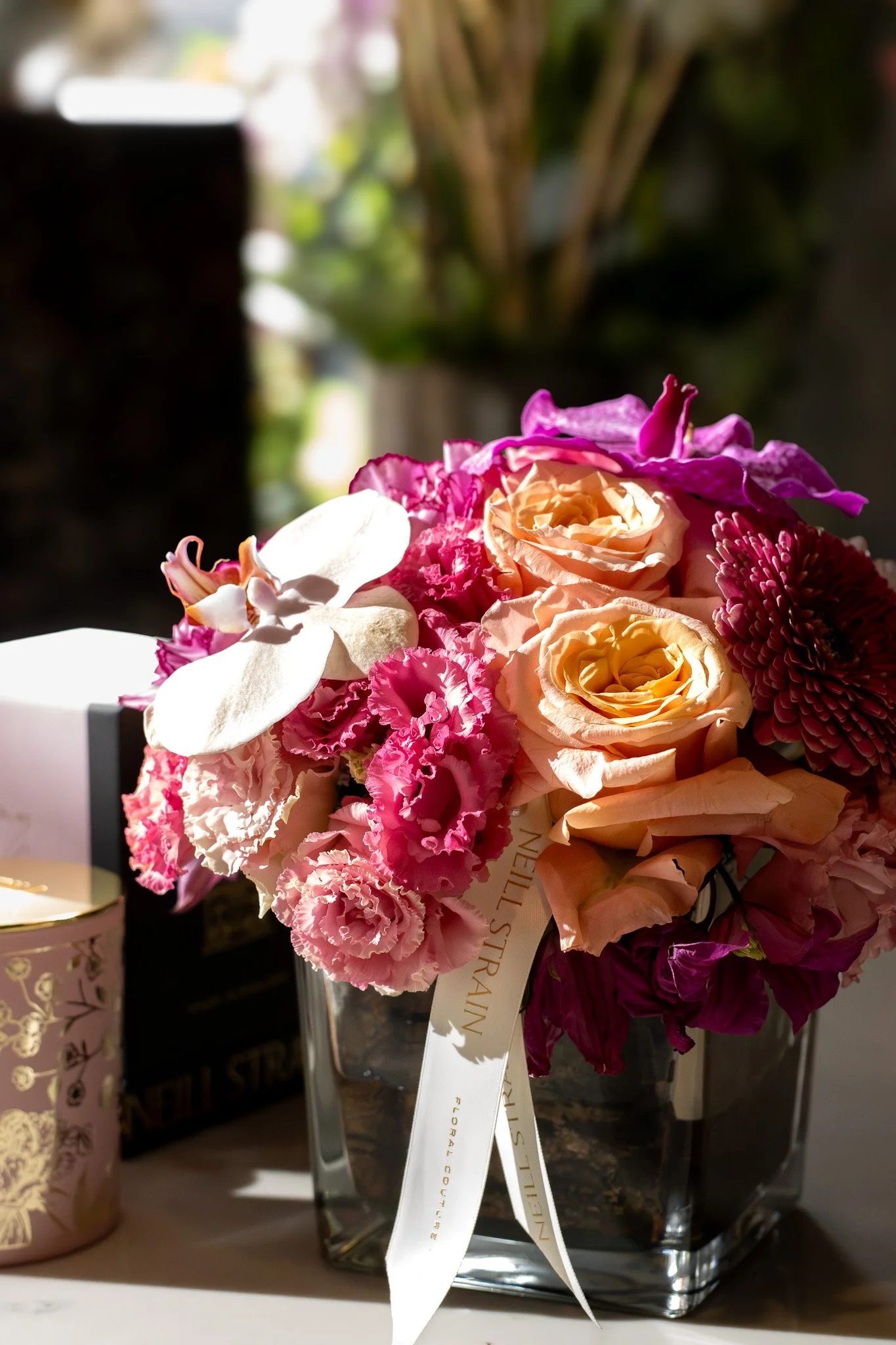 A colorful bouquet of flowers, including roses and other flowers, wrapped with a white ribbon, placed on a table with various decorative items and background greenery. Neill Strain, Harrods, Motcomb Street.