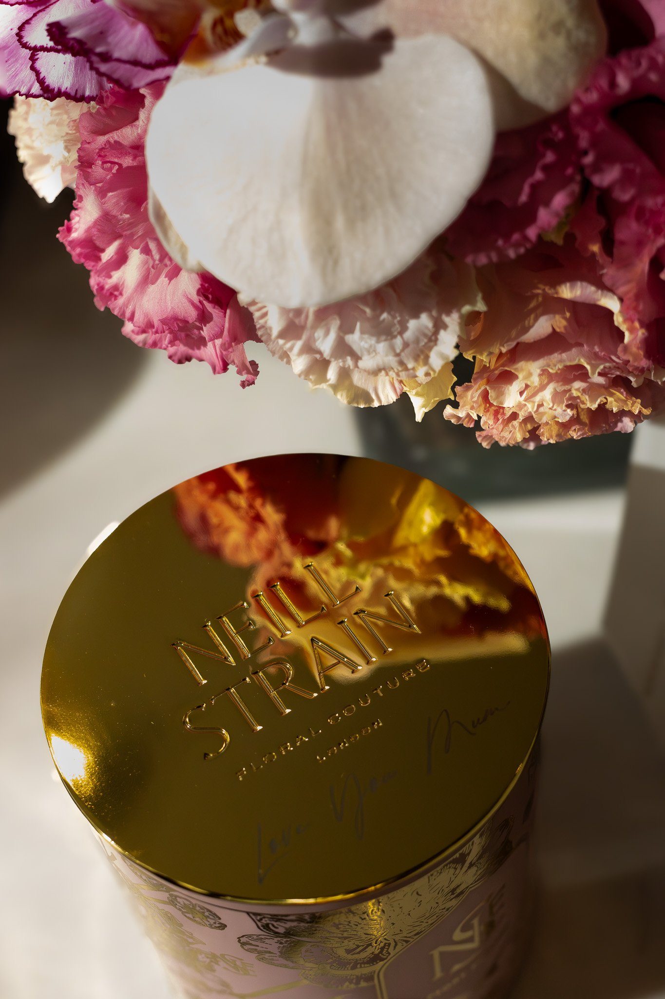 A close-up image of a gold-colored container with the text 'NEIL'S TRADING' engraved on top, surrounded by pink and white flowers. Neill Strain, Harrods, Motcomb Street.