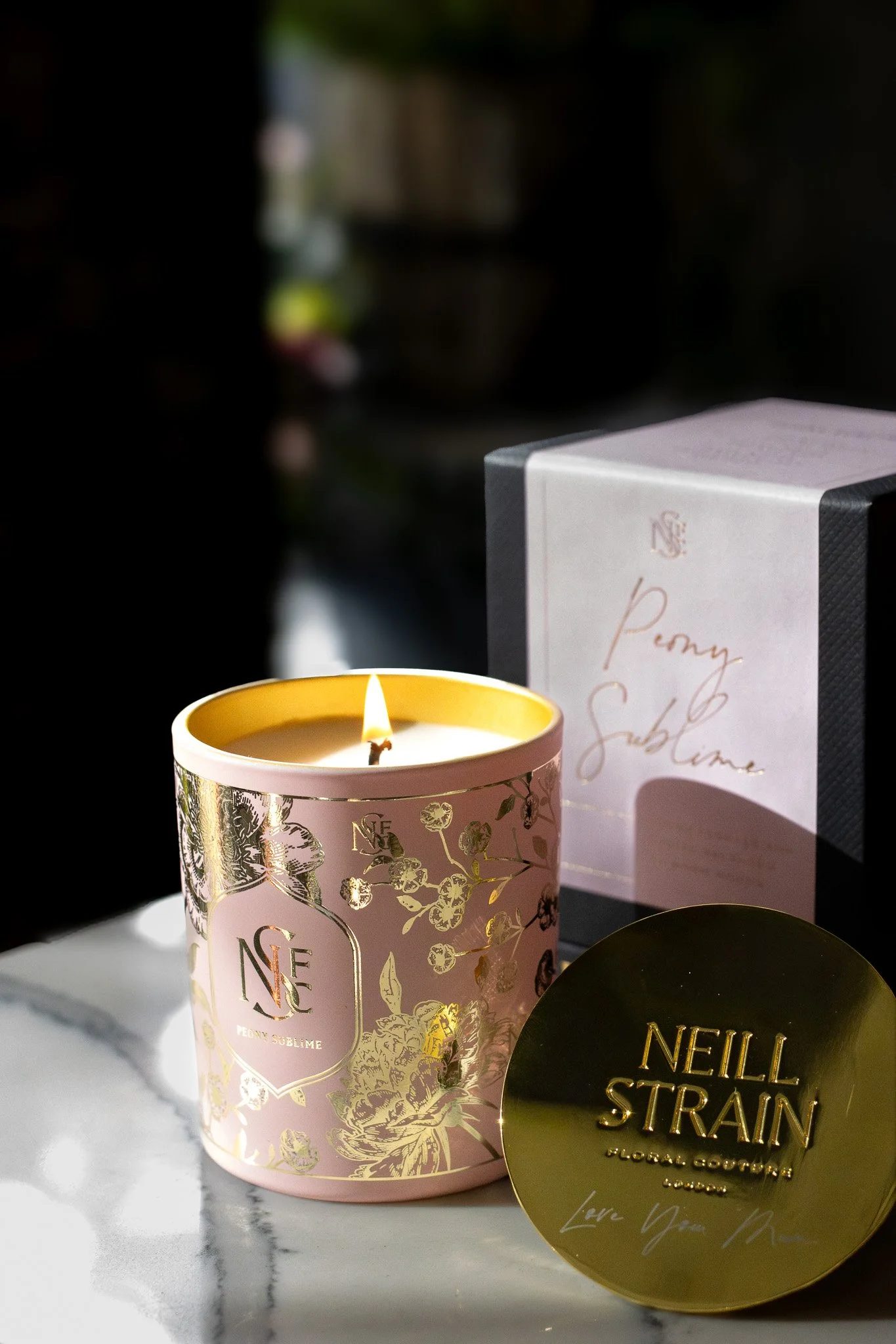 A pink and gold scented candle with floral design, a black and gold Neill Strain GG4 face mask package, and a pink box labeled 'Penny Sublime' on a white marble surface. Neill Strain, Harrods, Motcomb Street.