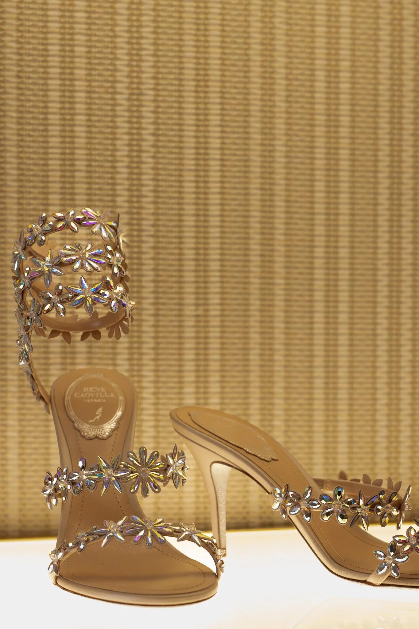 Beige high-heeled sandals decorated with flower-shaped rhinestones, displayed on a white surface with a textured golden background. Rene Caovilla.