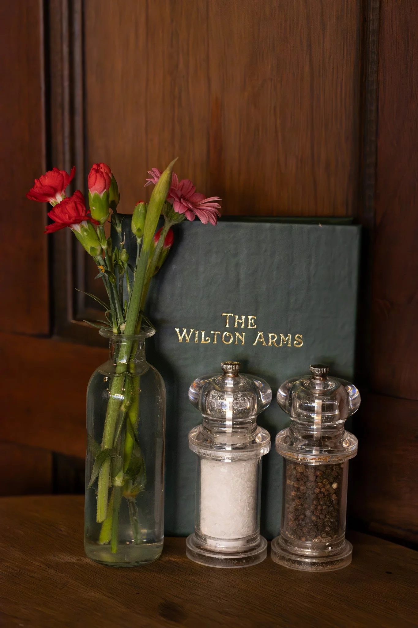 A vase of red and pink flowers, a green book titled 'The Wilton Arms', and three glass spice jars with white, black, and mixed spices on a wooden surface. The Wilton Arms.