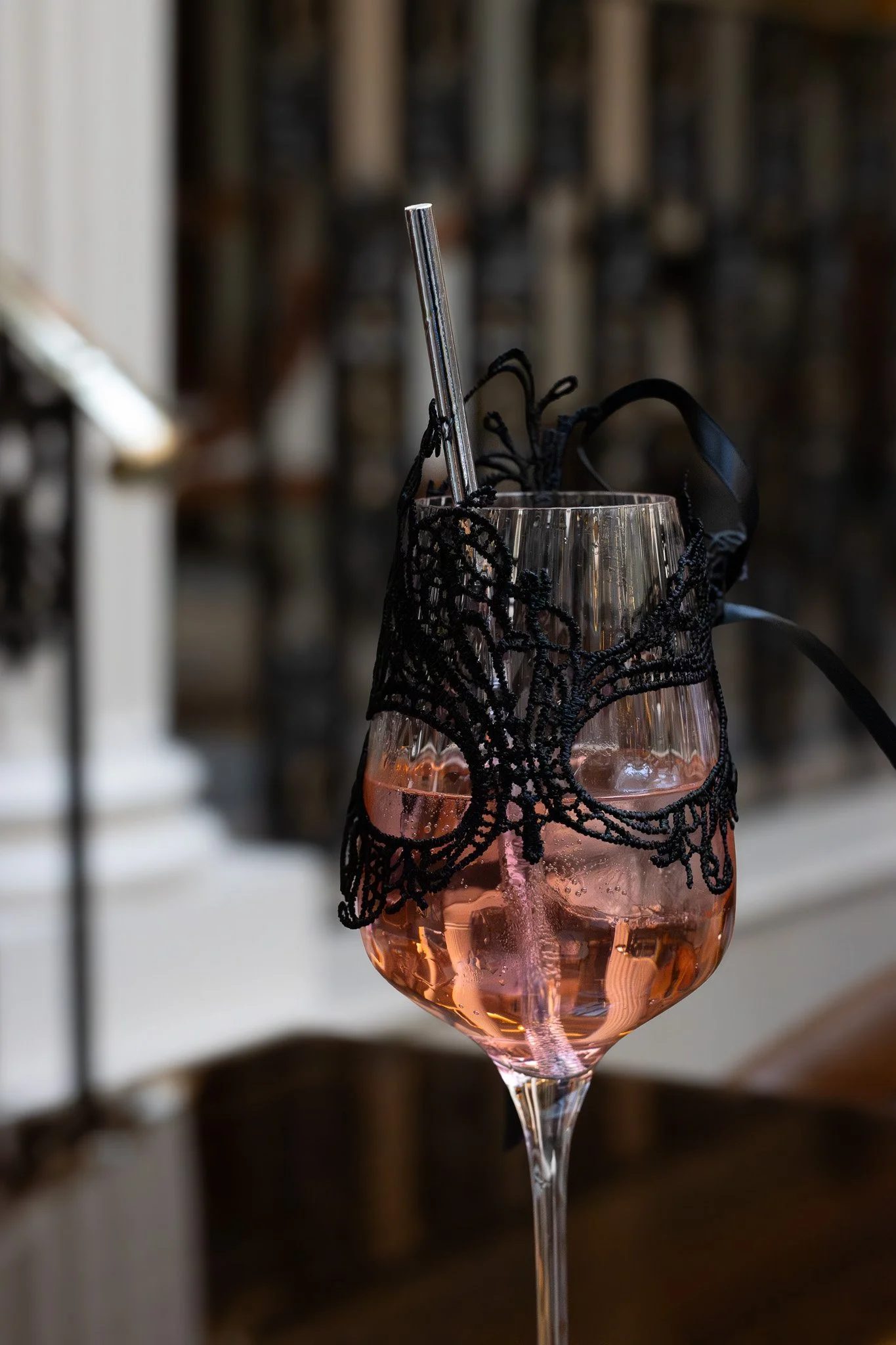 A glass of pink drink with black lace decoration around the rim, with a metal straw inside, set against a blurred background. The Lanesborough. Bridgerton Afternoon Tea.