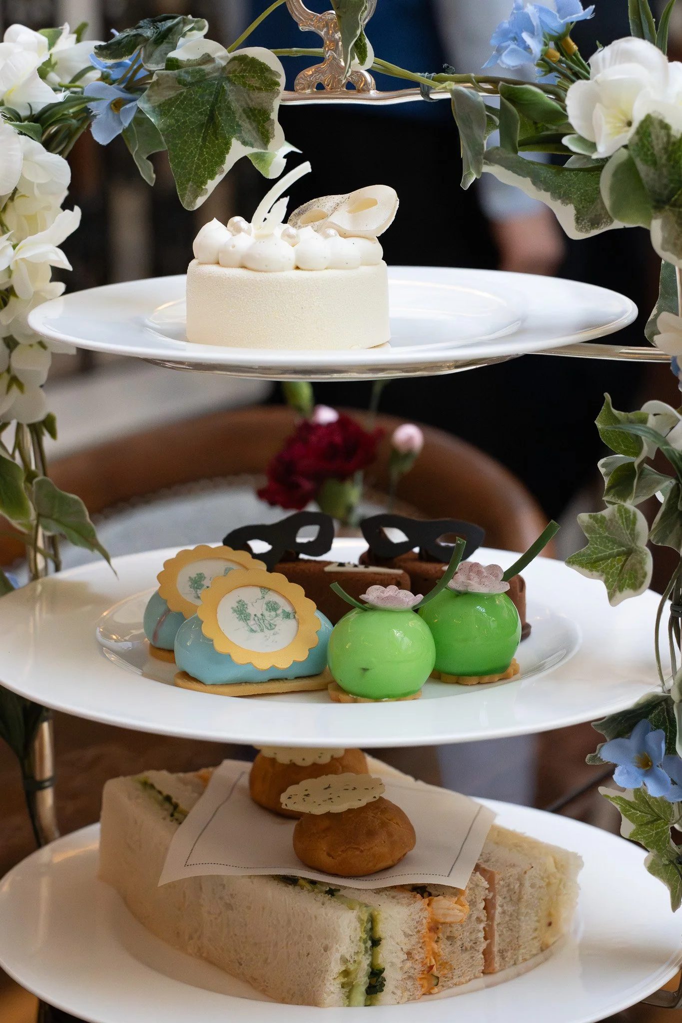 Bridgerton Afternoon Tea, with assorted desserts, including a white cake on the top, decorated with white frosting dollops and a white chocolate leaf, adorned with a floral arrangement of green leaves, white and blue flowers. The Lanesborough.