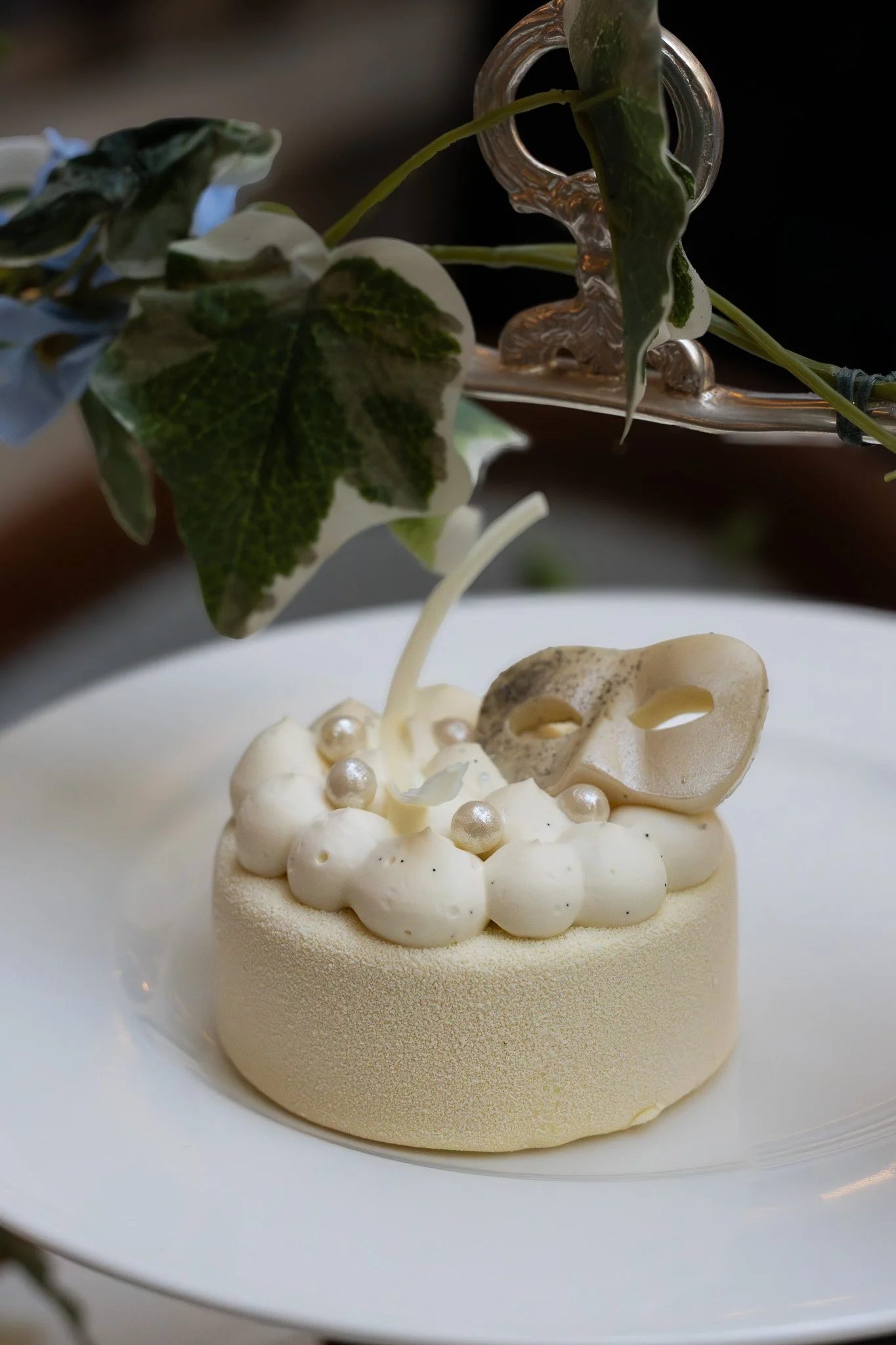 A white, round dessert topped with white cream or mousse, decorated with small edible pearls. It is served on a white plate, with green leaves and a silver decorative item in the background. The Lanesborough.