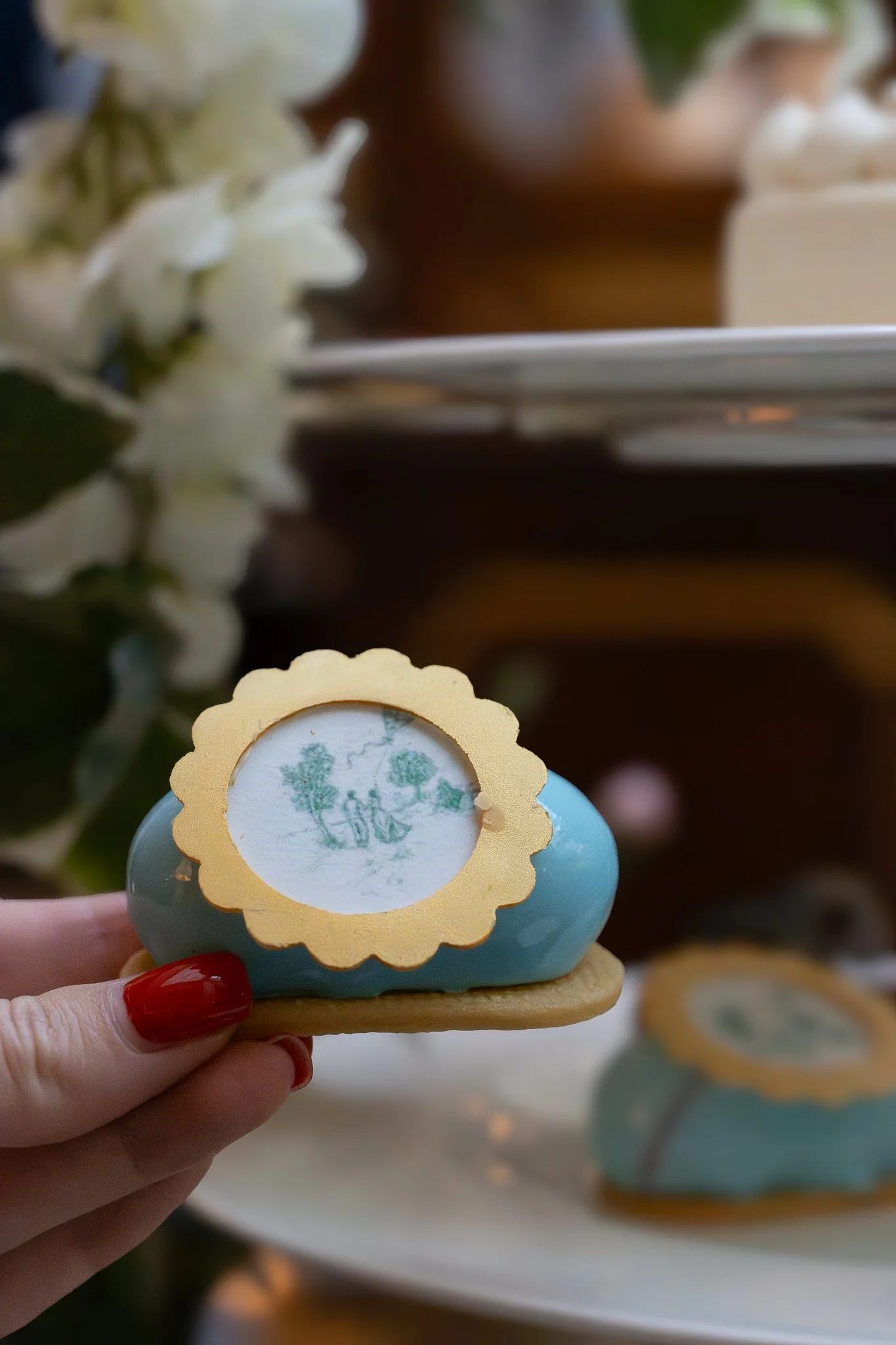 A hand with red nail polish holding a decorative blue Easter egg with a yellow scalloped edge and a white paper insert showing a printed image of a tree and people, in front of a table with more similar decorated eggs. The Lanesborough.