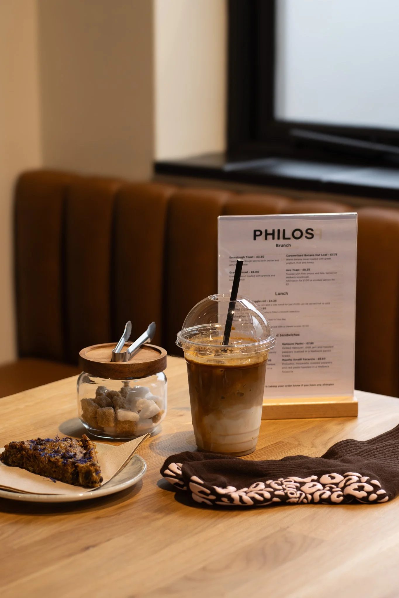 A table with a caramel frappuccino in a clear plastic cup with whipped cream on top, a small glass jar with sugar cubes, a slice of chocolate cake, a printed menu, and a folded cloth napkin with a black and white pattern. In the background, there is